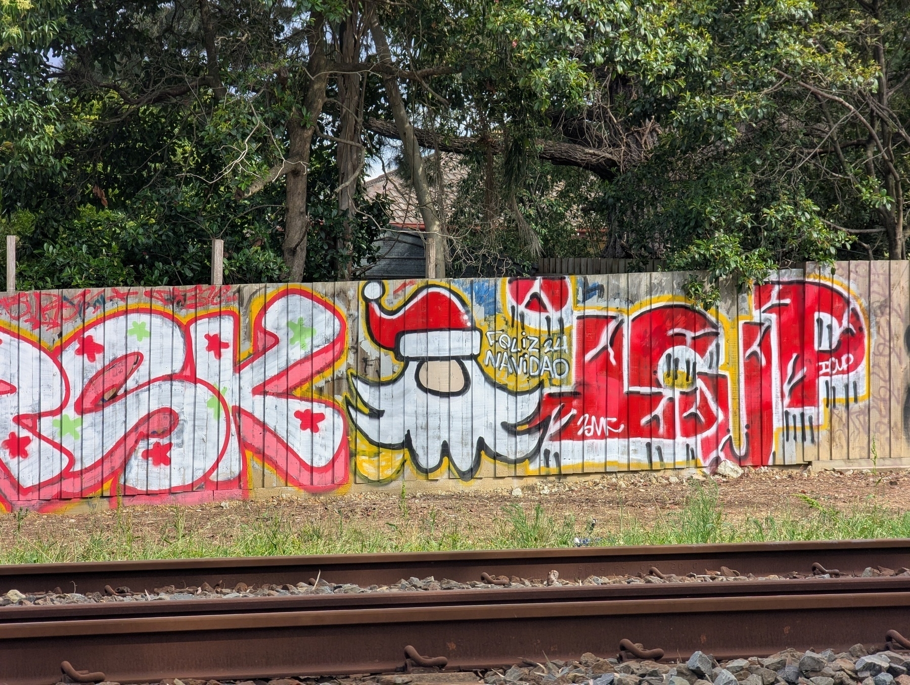 A colorful graffiti mural is painted on a wooden fence near train tracks, featuring Father Christmas and the message Feliz Navidad 24 amongst other abstract designs and text.