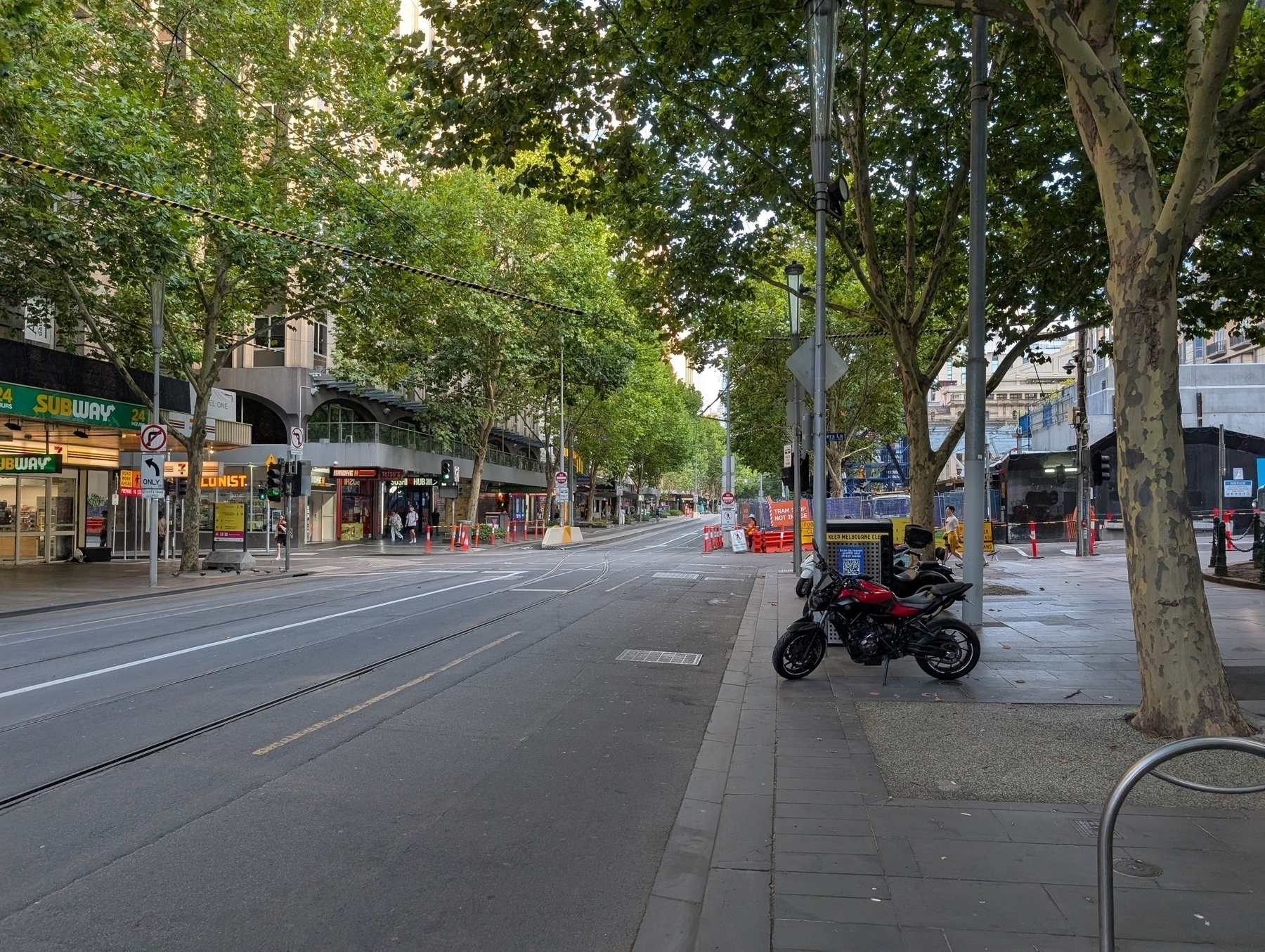 A quiet, tree-lined urban street features a couple of motorbikes parked on the sidewalk and several storefronts, including a Subway.