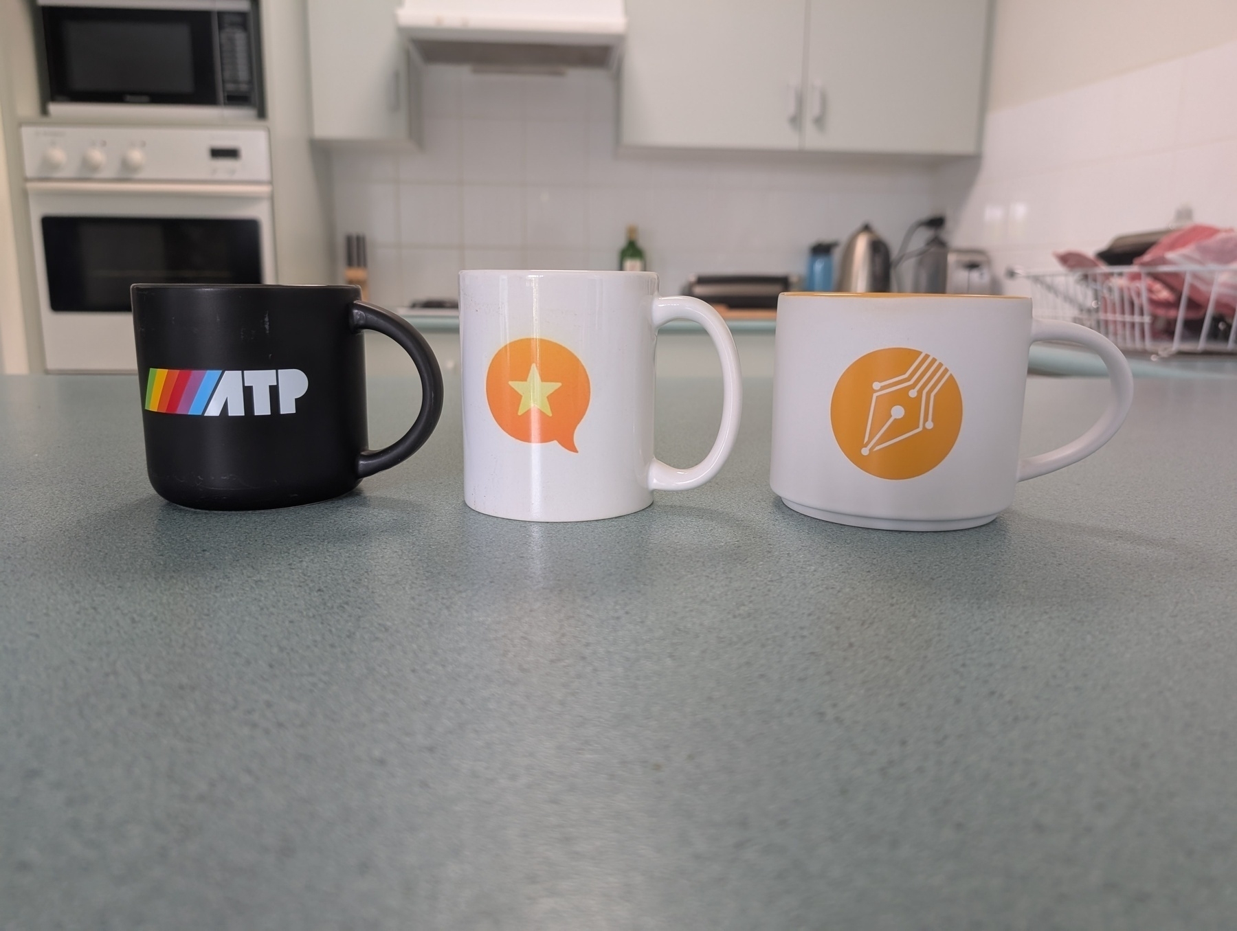 Three mugs with different designs — from left to right, the ATP logo, the Micro.blog logo, and the Stratechery logo — are lined up on a kitchen bench.