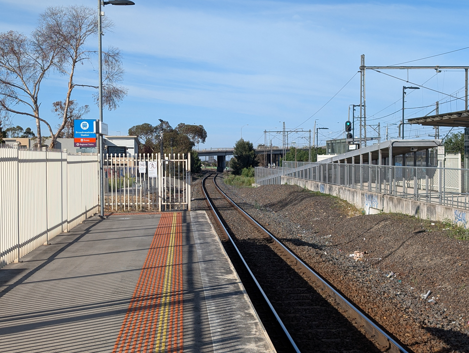 A railway platform with visible tracks, a fence, and overhead power lines stretches into the distance under a clear sky.