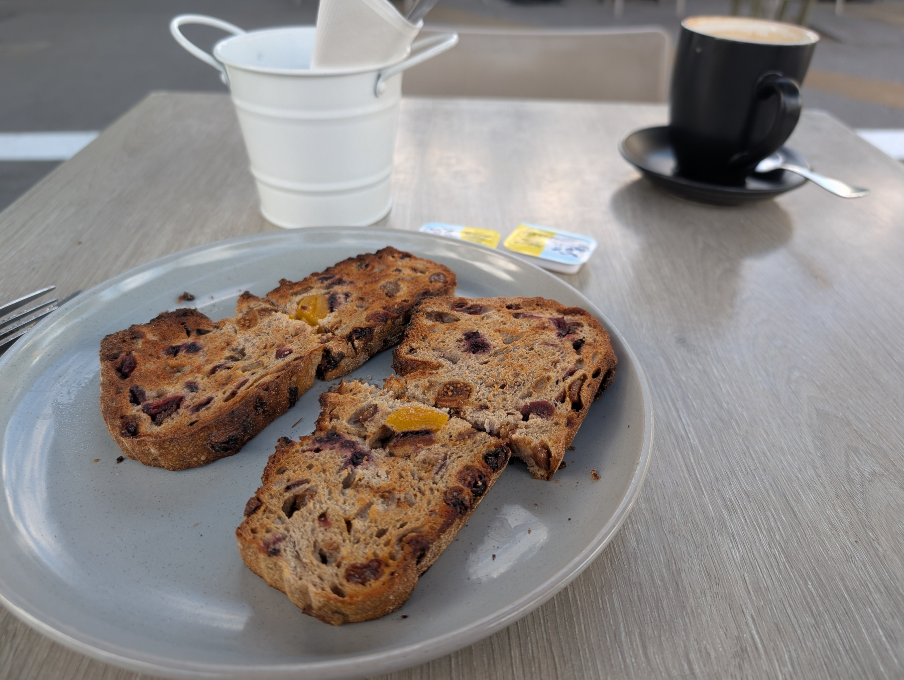 Two slices of fruit toast are on a plate next to a small bucket, with a coffee cup and butter packets in the background.