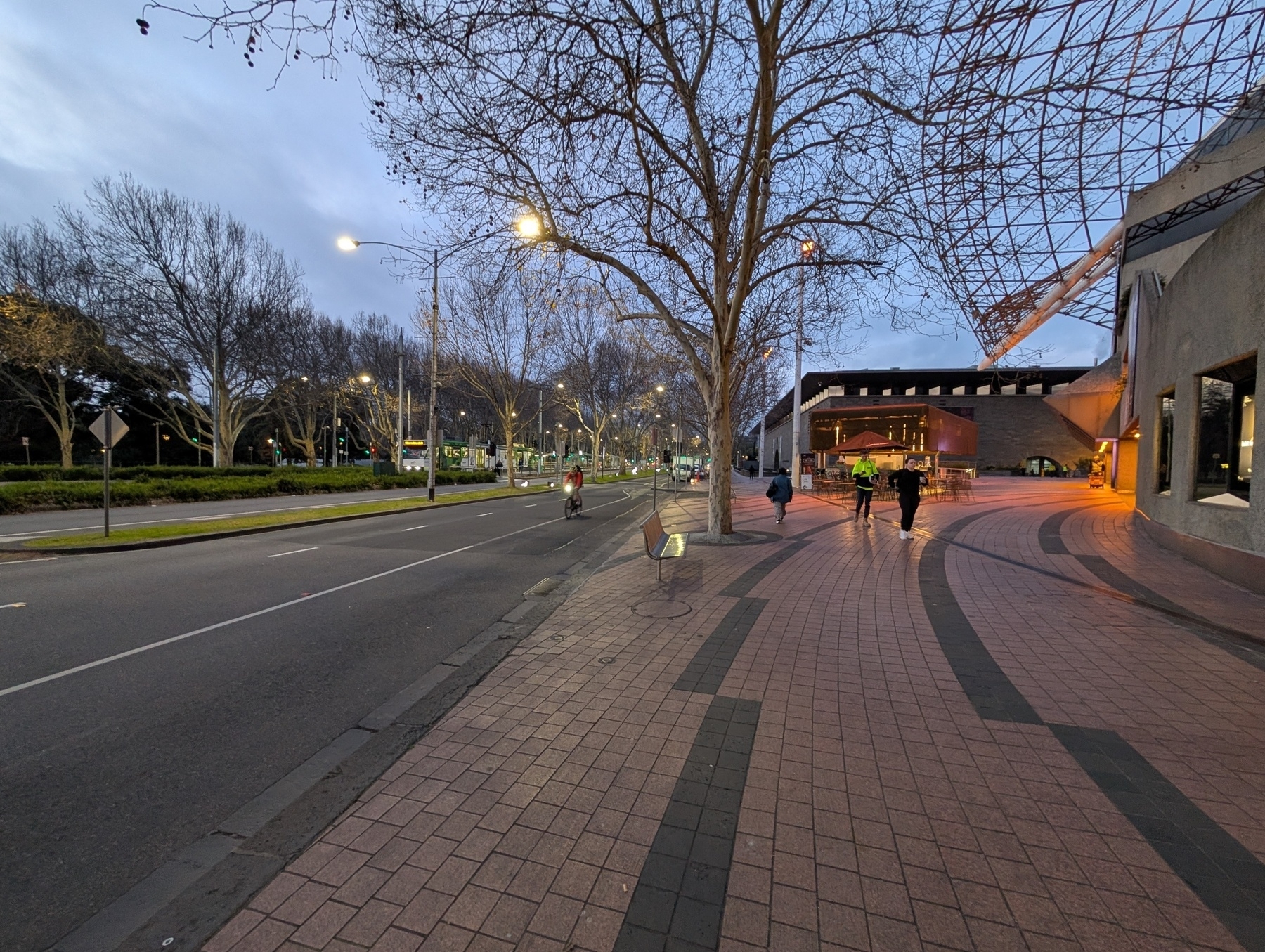 Auto-generated description: A paved walkway with a few people leads past a modern building under an overcast sky, with bare trees lining the adjacent street.