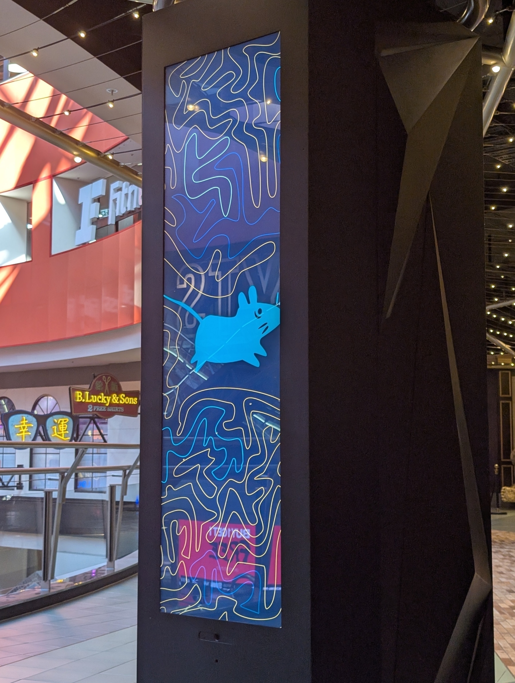 A tall digital display in a shopping centre shows an abstract blue and purple design with a mouse silhouette, surrounded by various shops.