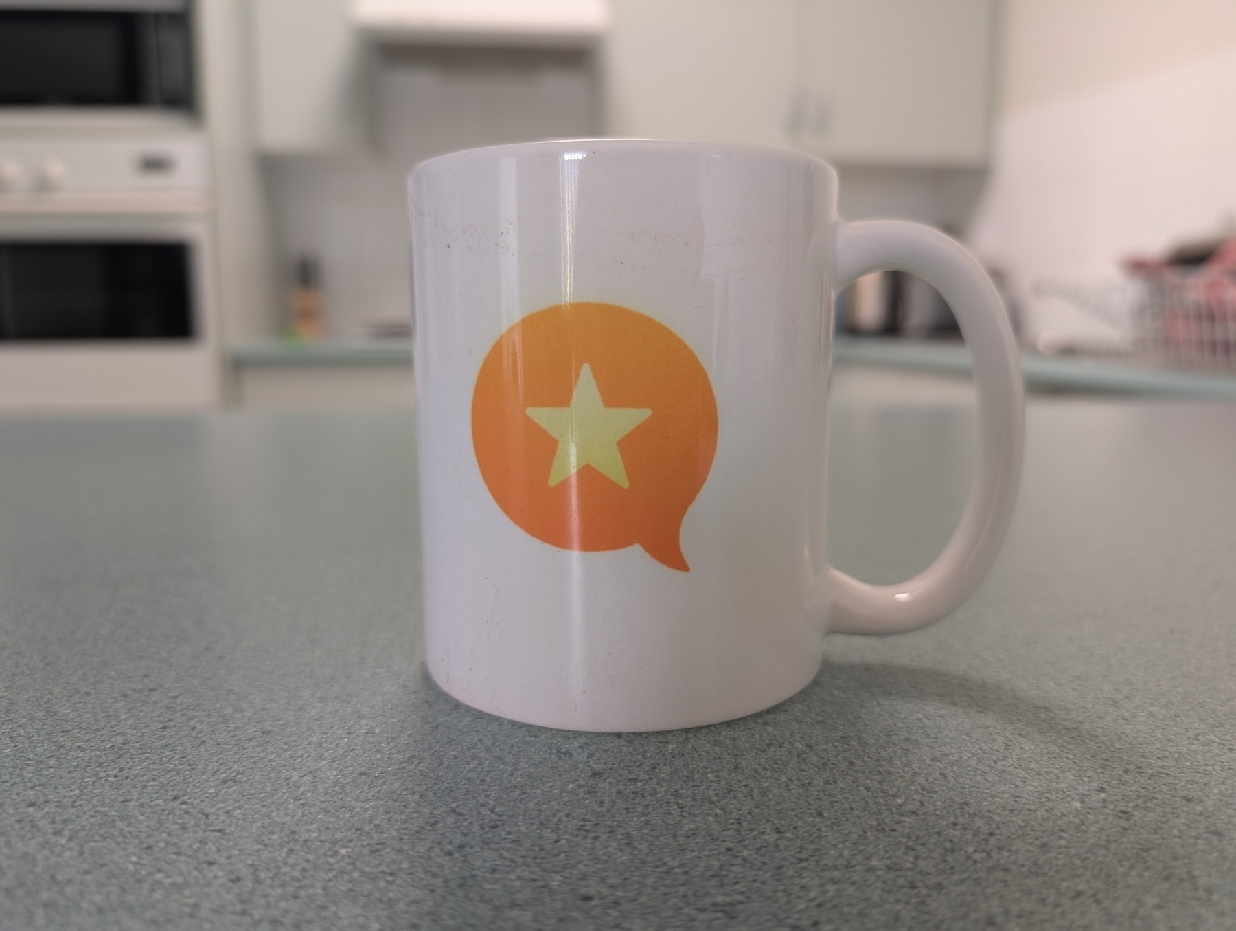 A white mug with the Micro.blog logo — an orange speech bubble design featuring a yellow star — is placed on a kitchen bench.