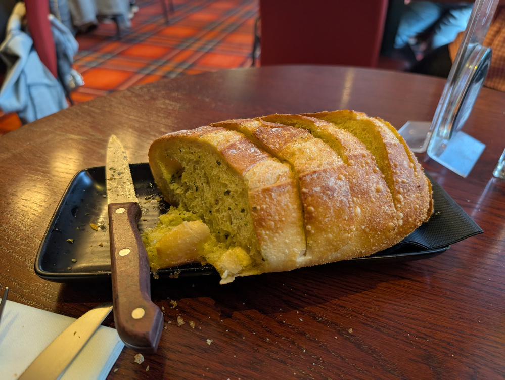 A sliced loaf of bread is placed on a black rectangular plate beside a wooden-handled knife on a wooden table.