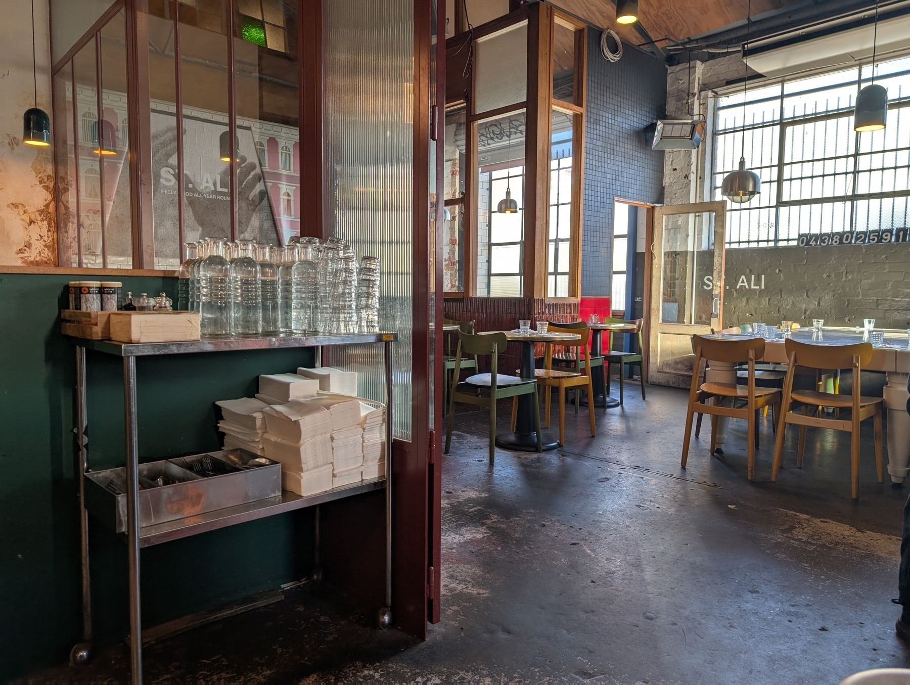 A cozy restaurant interior features wooden tables and chairs, with a corner shelf stocked with bottled water and takeout containers.