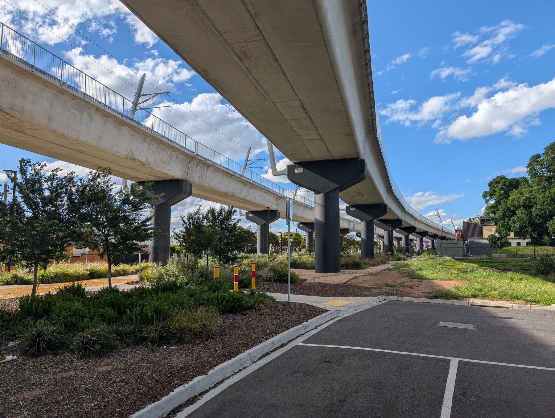 Auto-generated description: A concrete overpass runs parallel to a paved pathway and landscaped area, all beneath a blue sky with scattered clouds.