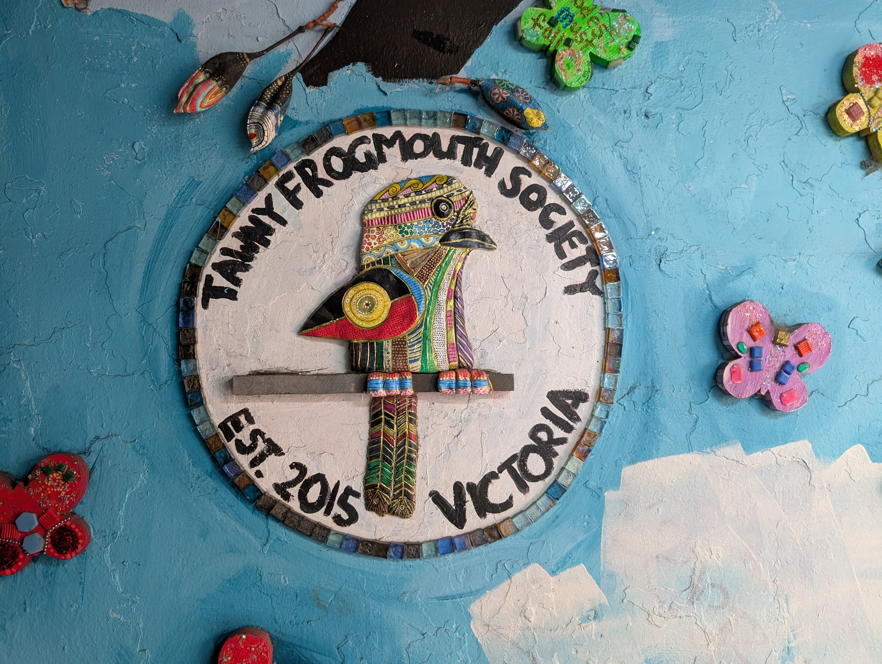 A colorful mosaic featuring a bird with the text Tawny Frogmouth Society Est. 2015 Victoria surrounded by artistic floral designs on a blue background.