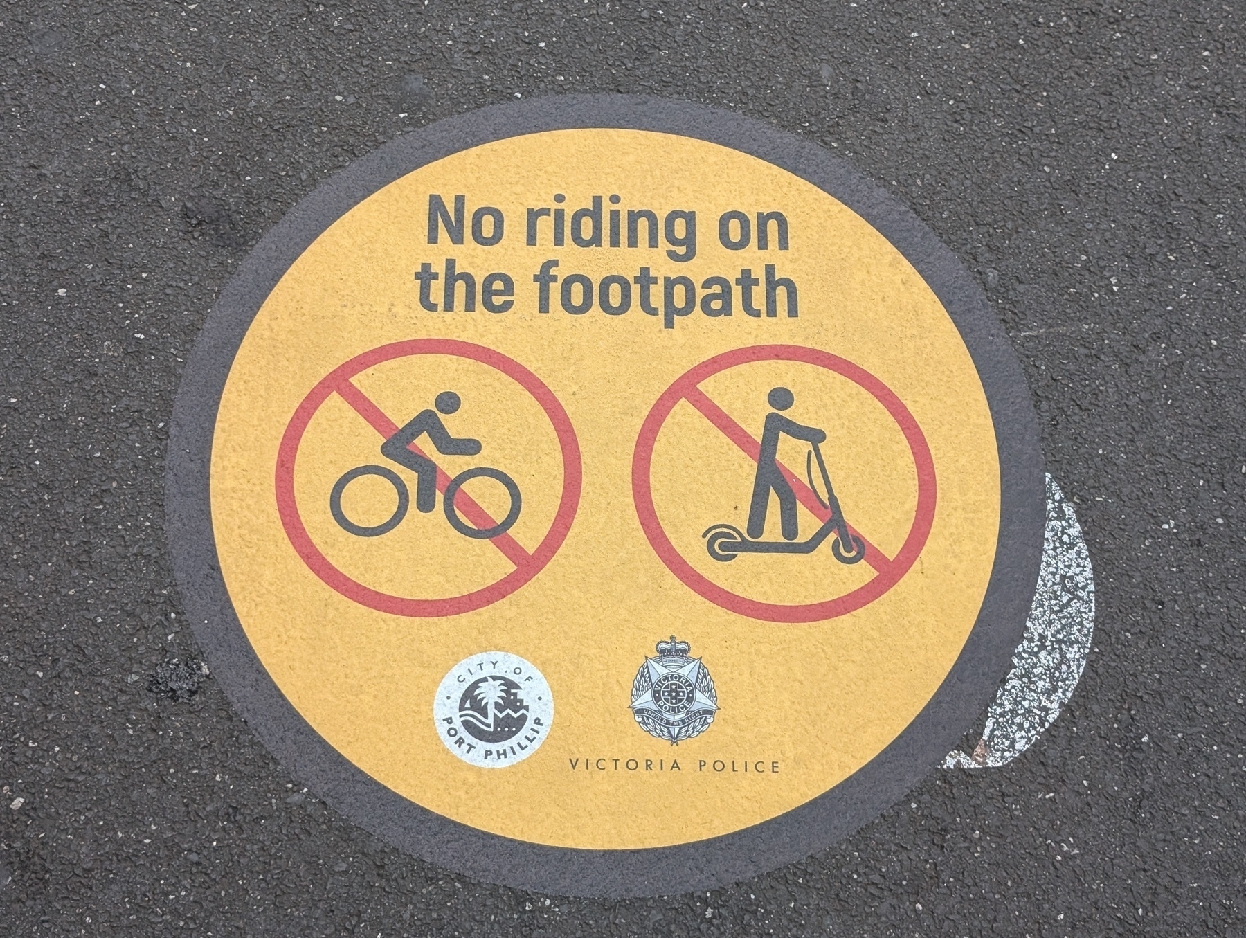 A circular sign on the ground prohibits riding bicycles and scooters on the footpath, featuring symbols of both with red slashes through them.