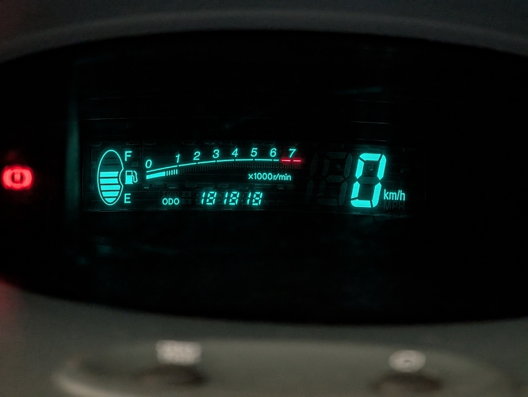 A car dashboard displays a digital speedometer reading 0 km/h, an odometer at 181818, and a fuel gauge indicating moderate amount of fuel.