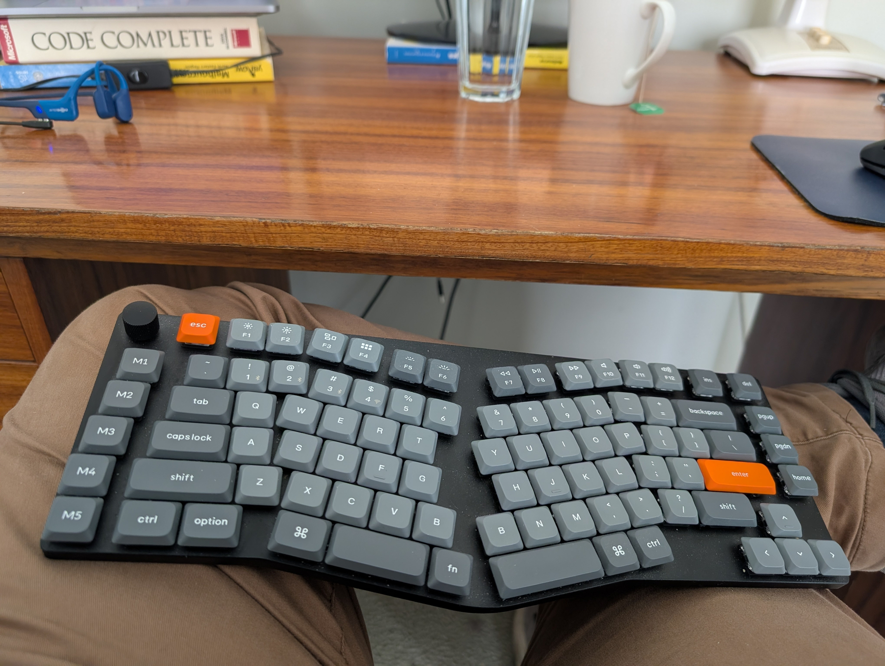 A split mechanical keyboard resting on a persons crossed leg with an orange enter key, sitting at a wooden desk with programming books and a coffee mug.