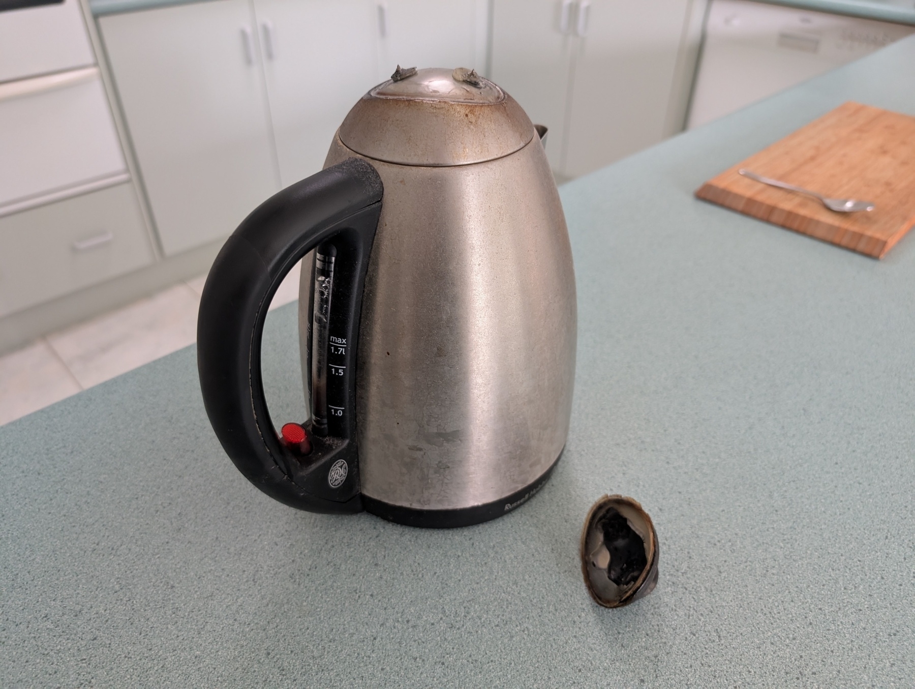 Auto-generated description: A metallic electric kettle with a detached lid is placed on a kitchen bench.