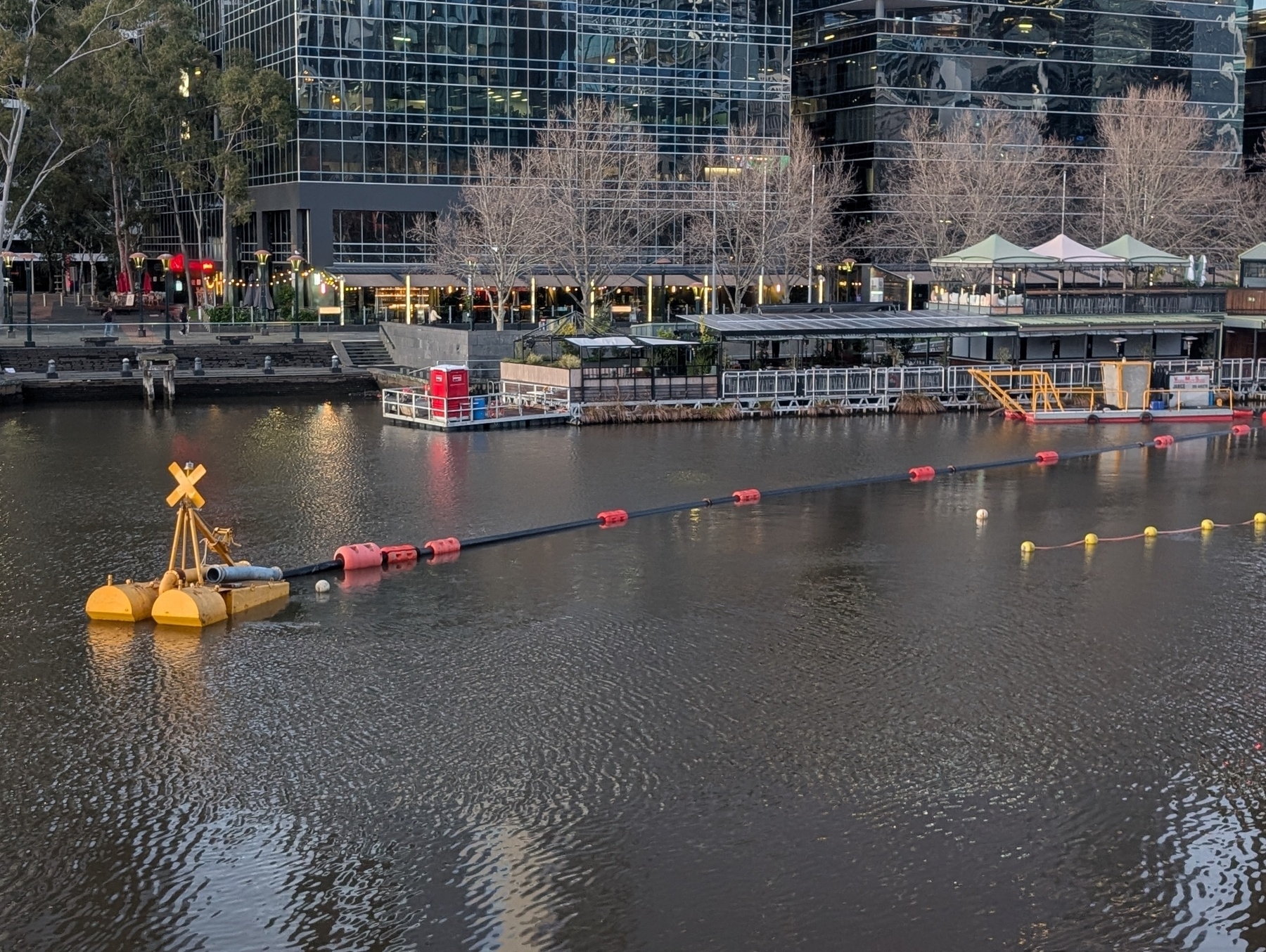 Auto-generated description: A floating barrier with red and yellow buoys stretches across a urban riverfront near modern glass buildings.