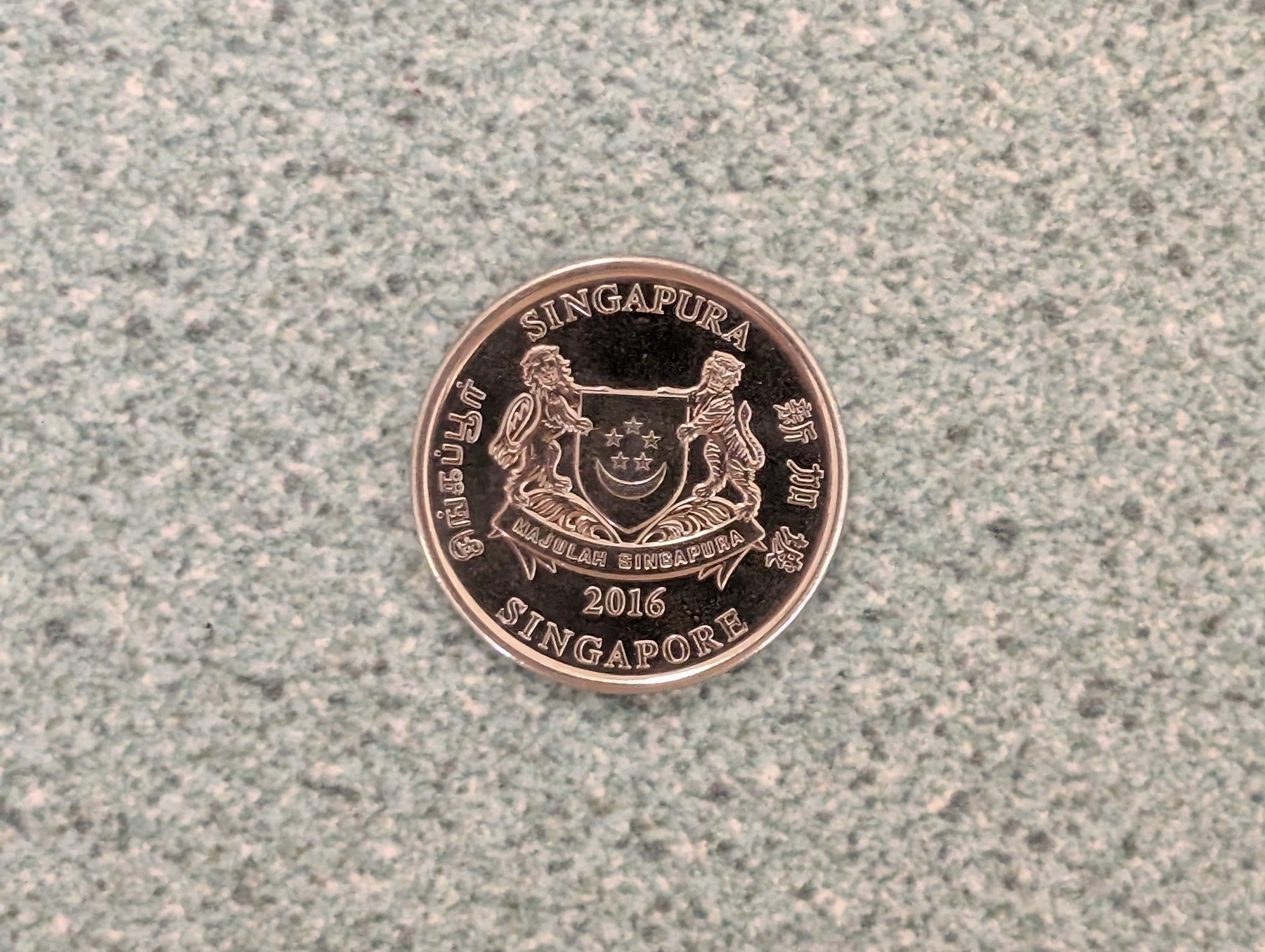 A coin from Singapore minted in 2016 is displayed on a textured surface, featuring the country's coat of arms and inscriptions in multiple languages.