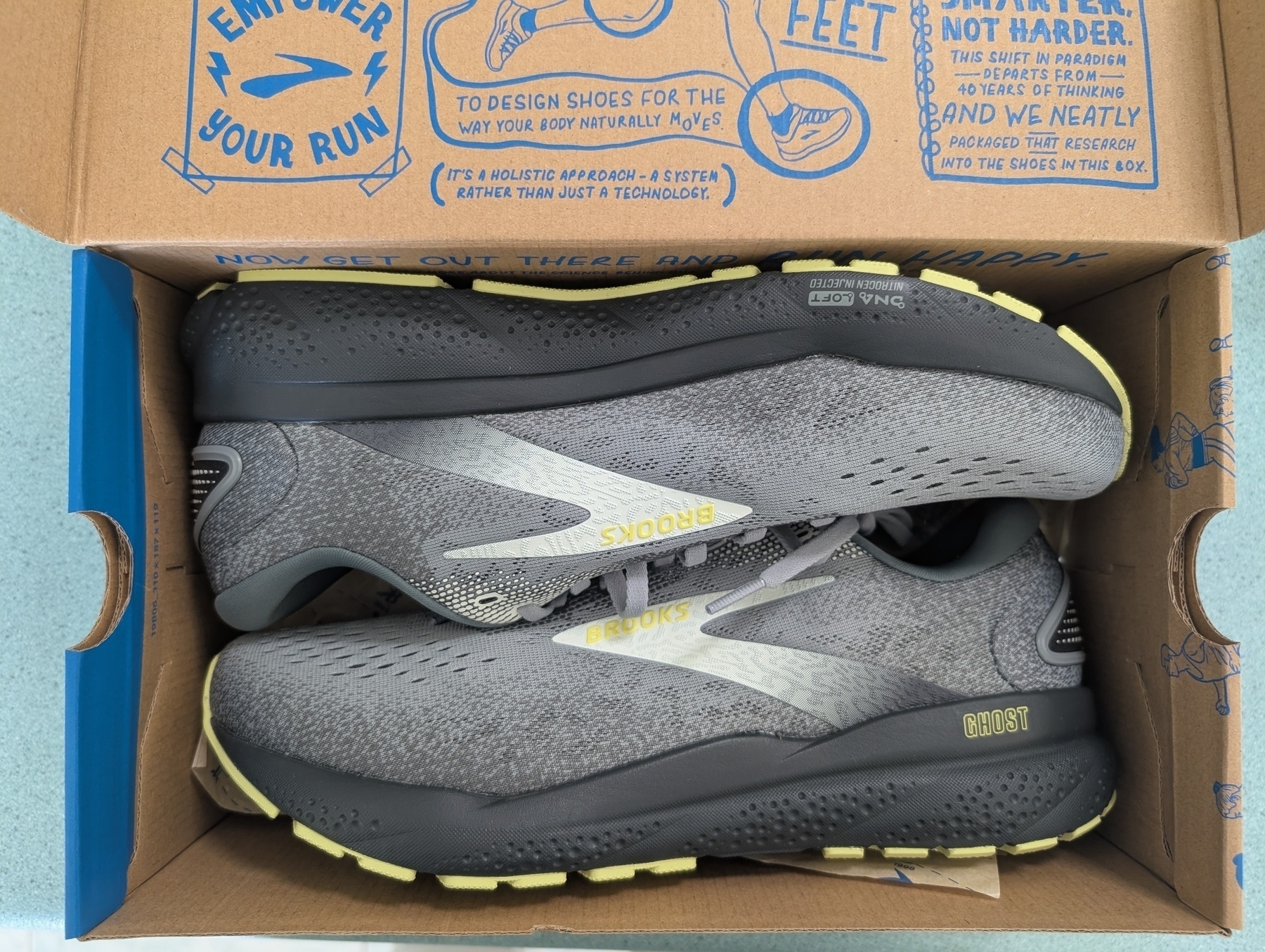 Auto-generated description: A pair of gray Brooks running shoes with yellow accents is displayed in an open shoebox with blue printed graphics and text on the inside lid.