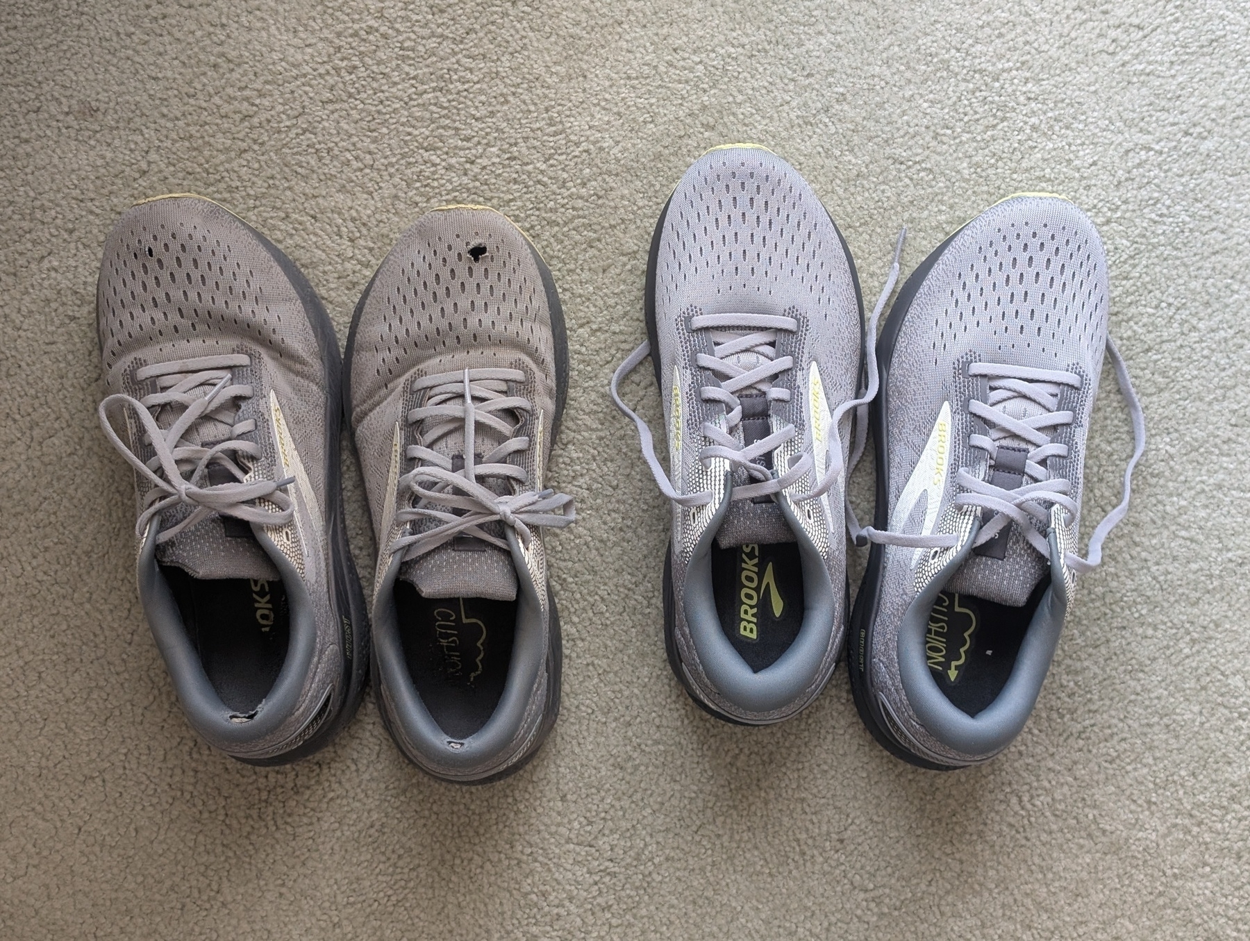 Auto-generated description: Two pairs of grey running shoes are placed side by side on a carpeted floor, with one pair appearing more worn than the other.