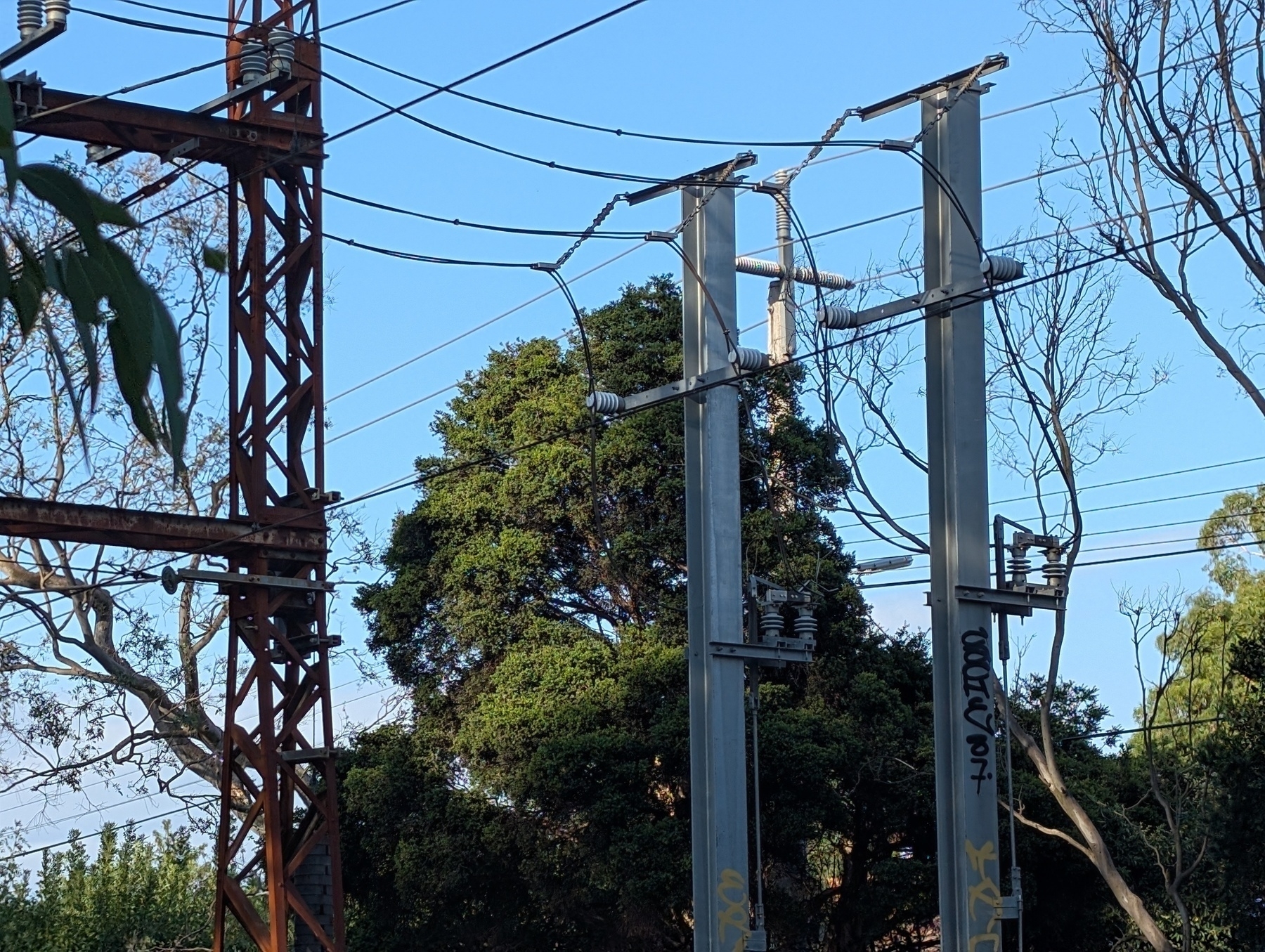 Auto-generated description: Power lines and electrical structures with closed isolator switches are set against a backdrop of trees and a clear blue sky.