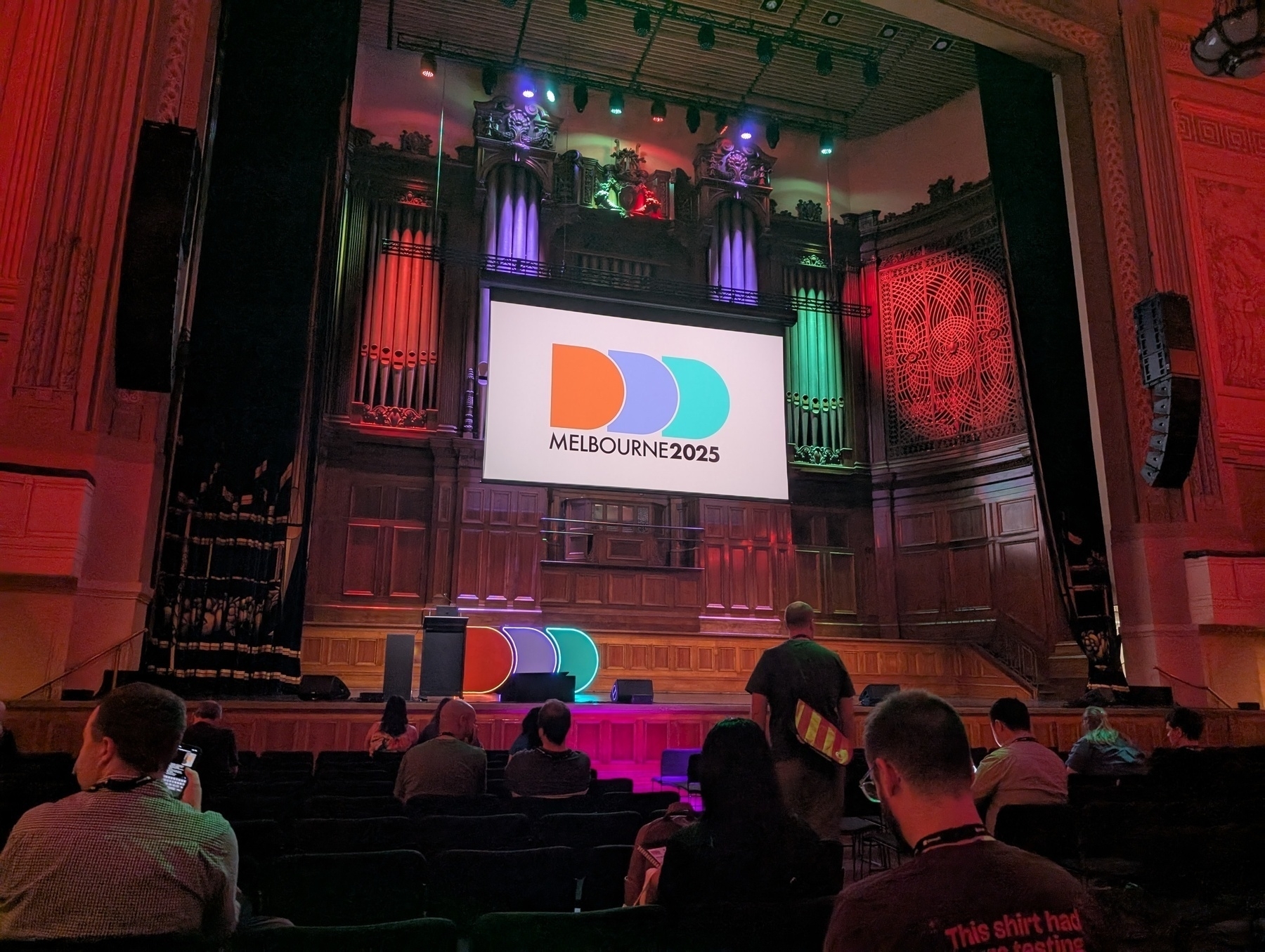 Auto-generated description: A stage featuring a Melbourne 2025 banner is set up in a theatre with people seated and colourful lighting.