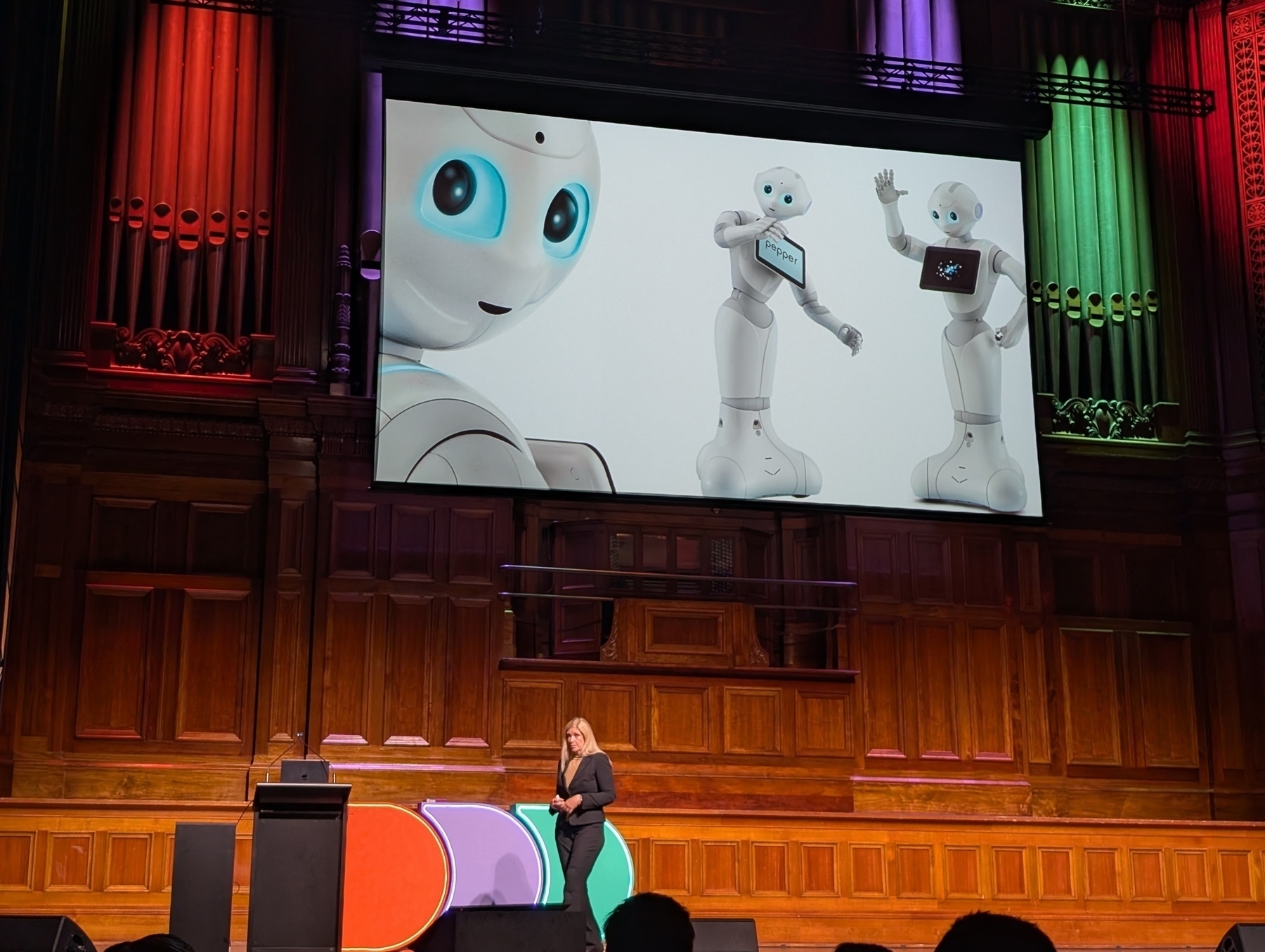 Auto-generated description: A speaker is presenting on stage with a slide displaying images of humanoid robots.