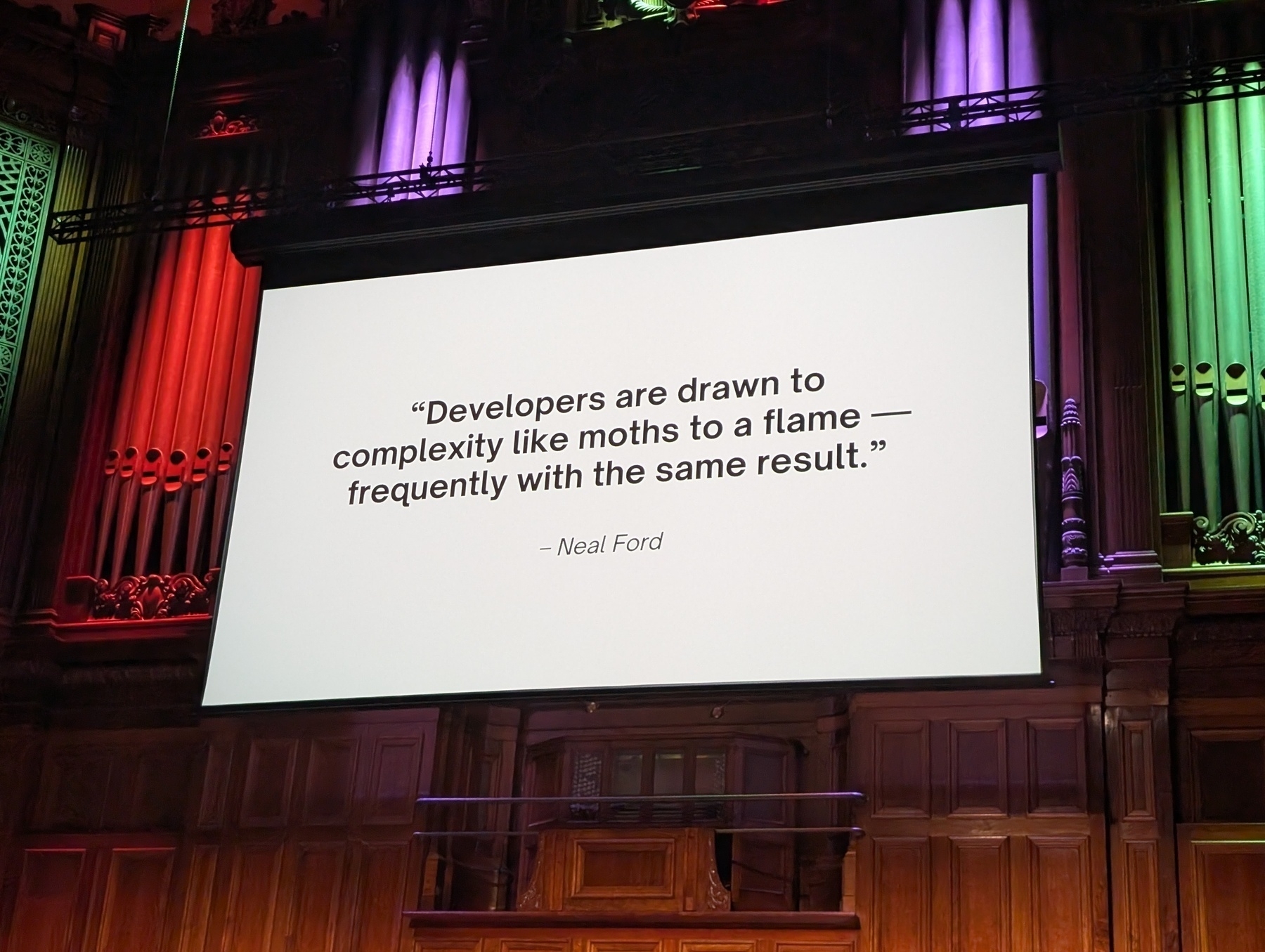 Auto-generated description: A large screen displays a quote by Neal Ford about developers and complexity, set in a grand hall with colorful lighting.