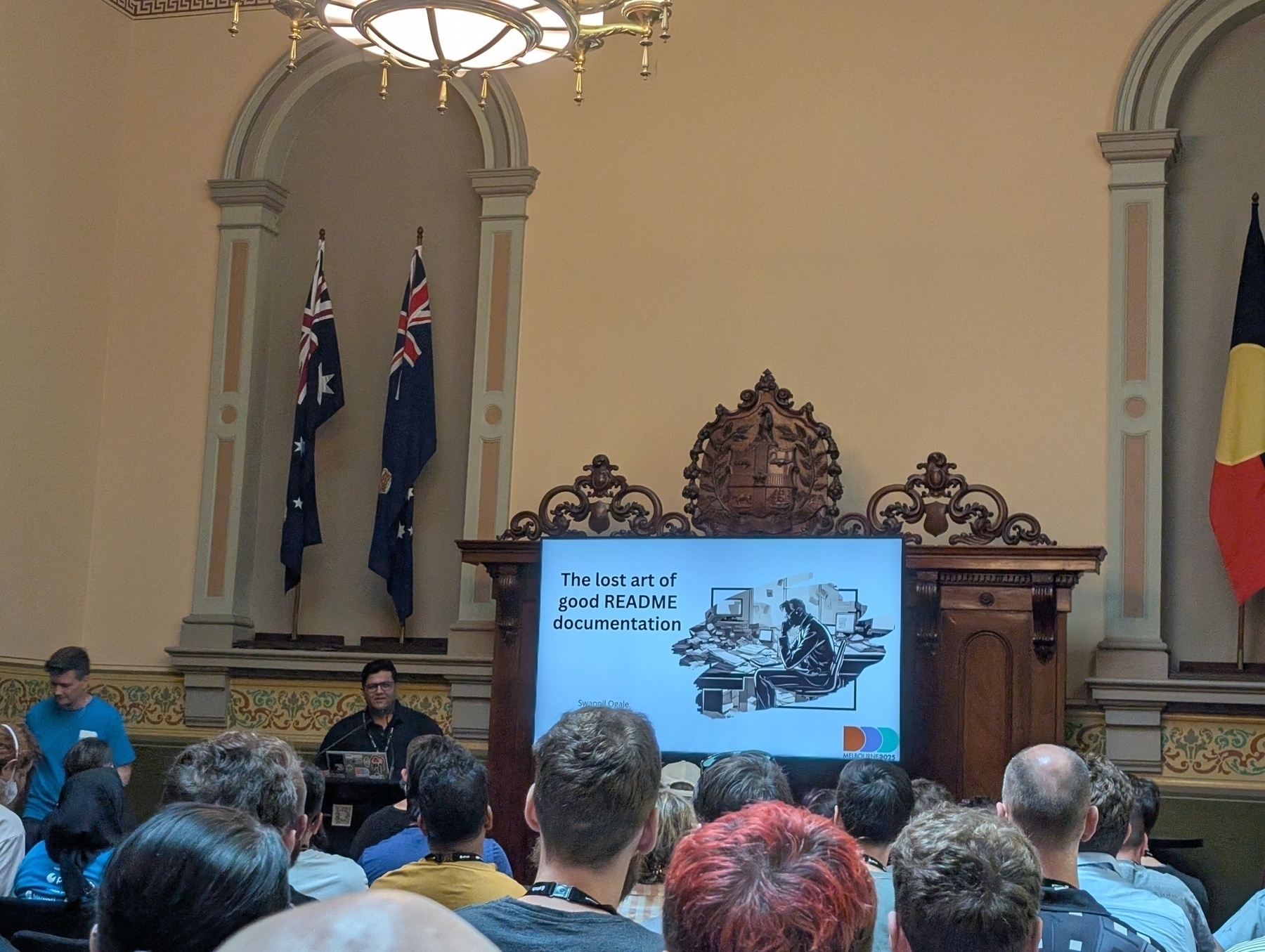 Auto-generated description: A speaker is presenting on The lost art of good README documentation to an audience in a formal room with flags and ornate decor.