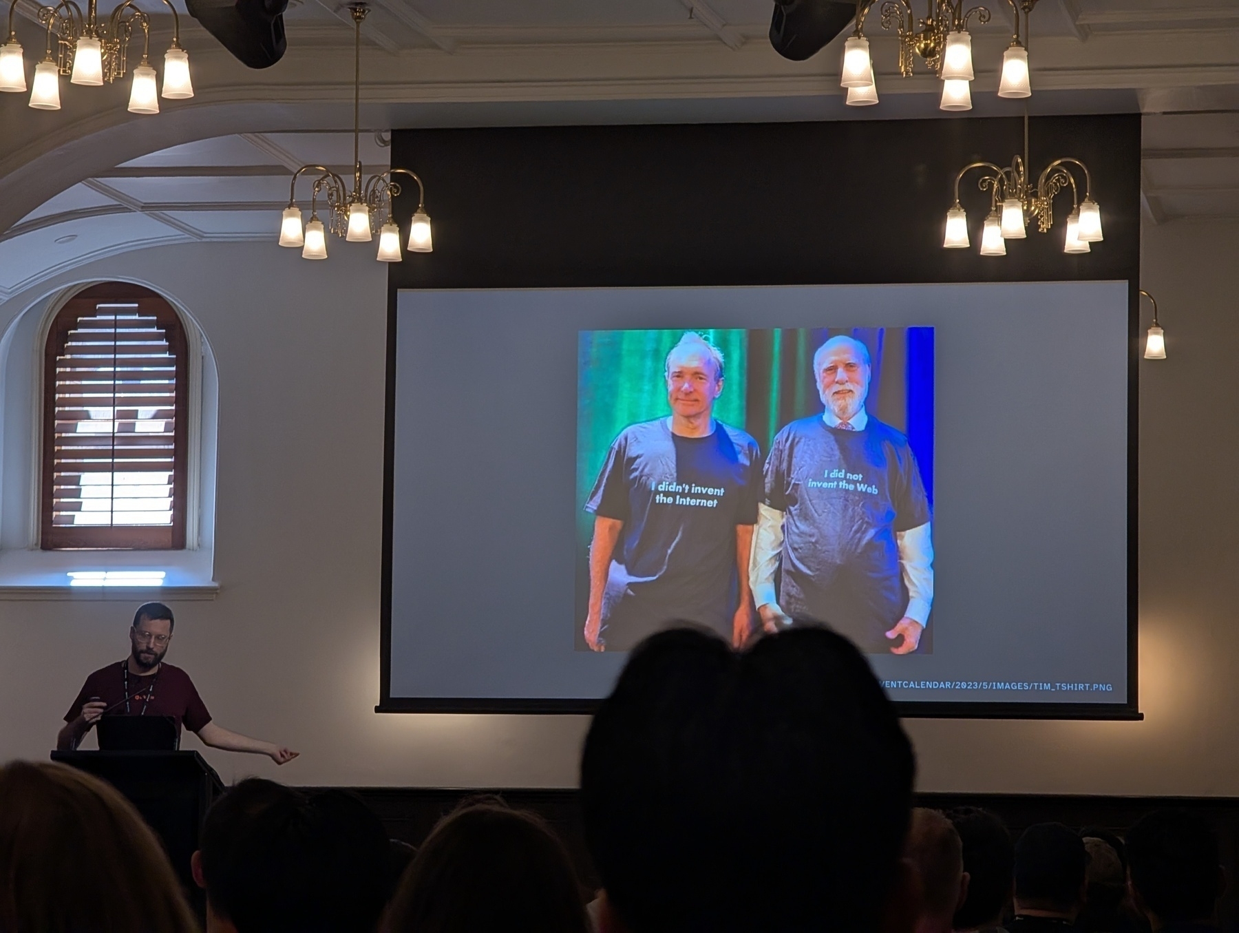 Auto-generated description: A presenter stands in front of an audience, showing a slide with two individuals wearing t-shirts that read I blame Immanuel for this behavior.