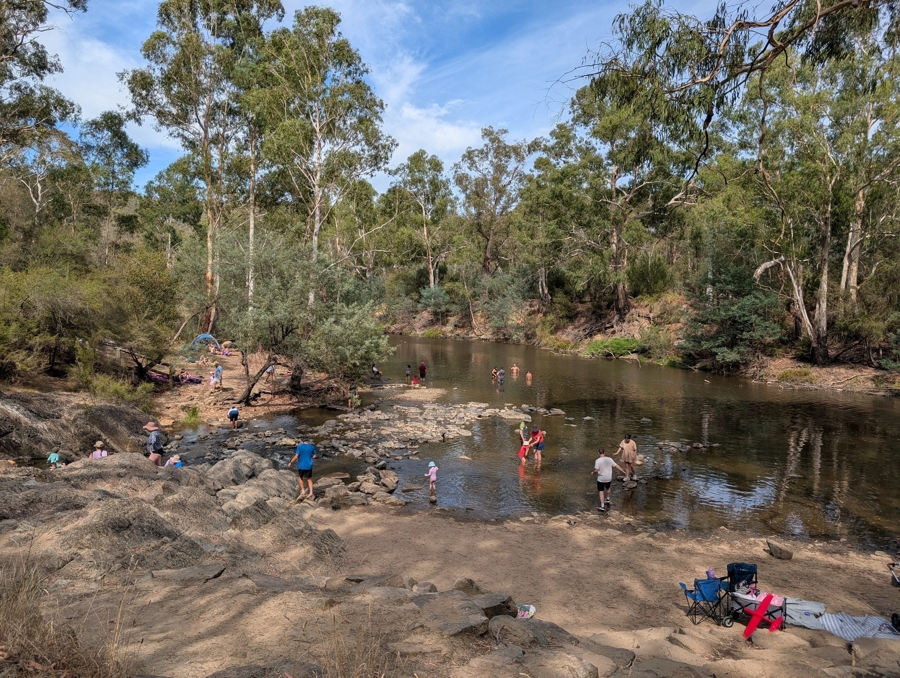 Auto-generated description: People are enjoying a day out by a river surrounded by trees and rocks, with some swimming and others relaxing on the shore.