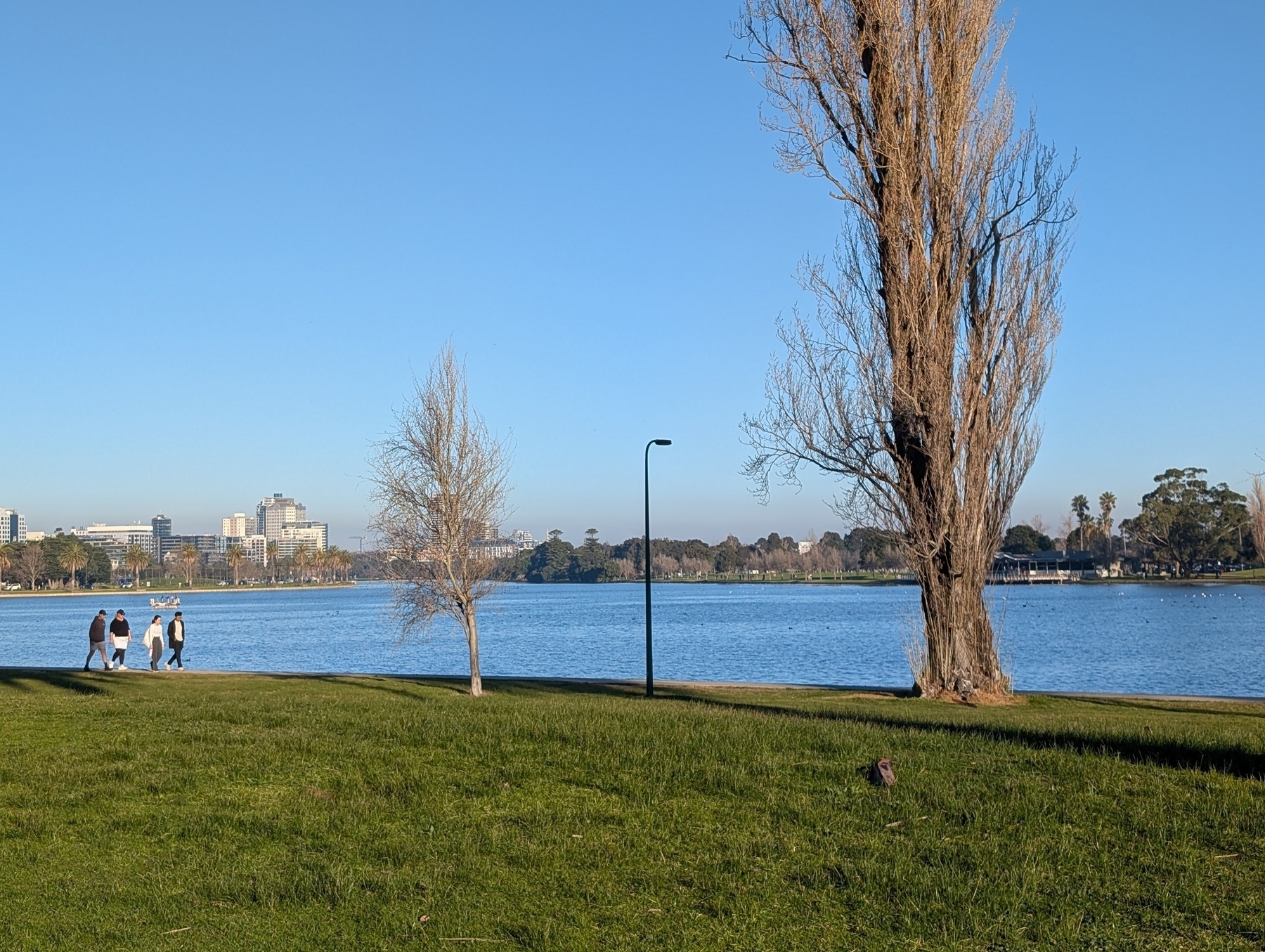 Auto-generated description: Three people walk by a lakeside park with grassy areas, trees, and a clear blue sky.