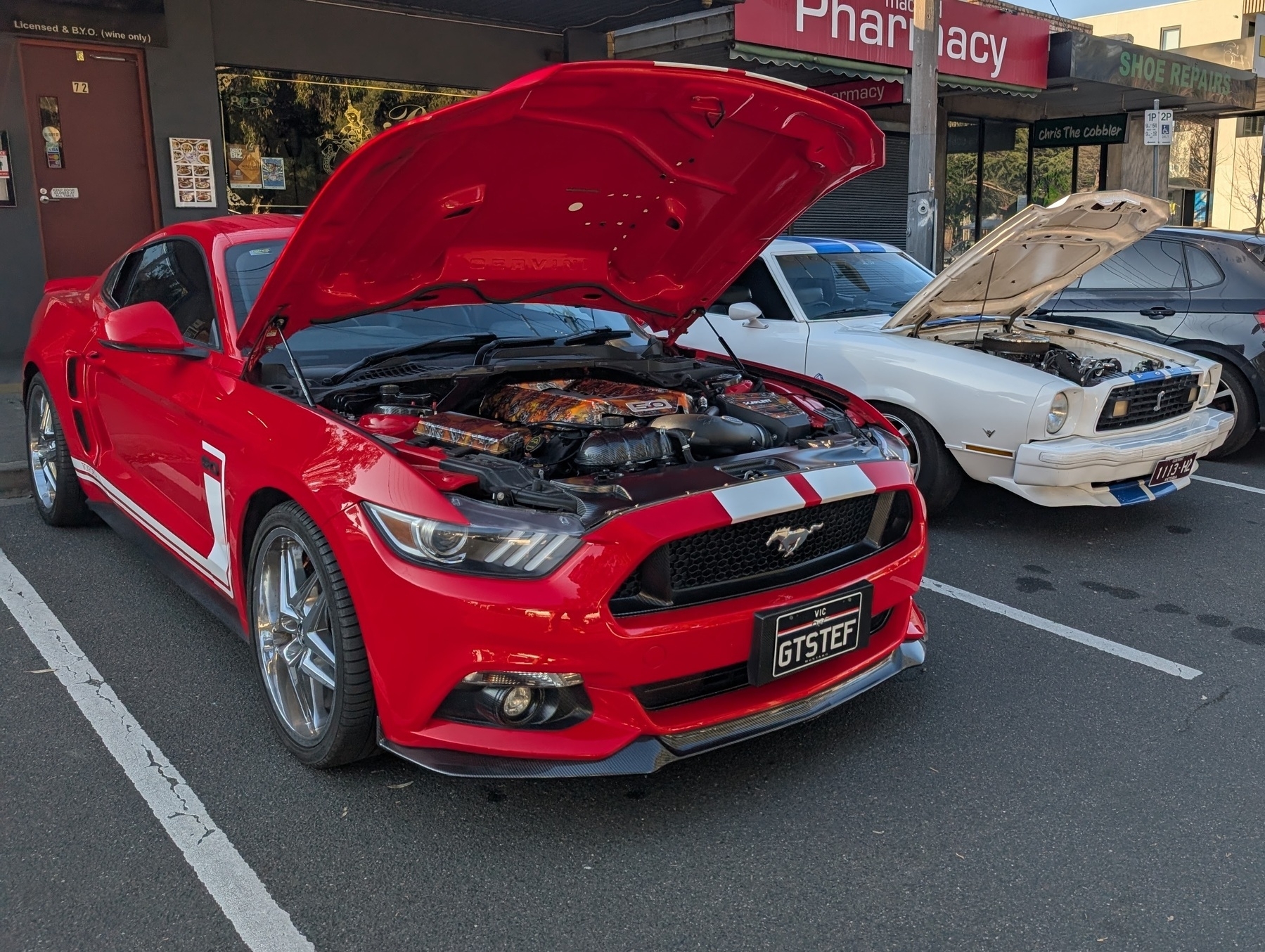 Auto-generated description: Two cars with open bonnets are parked on the street, one red and one white.