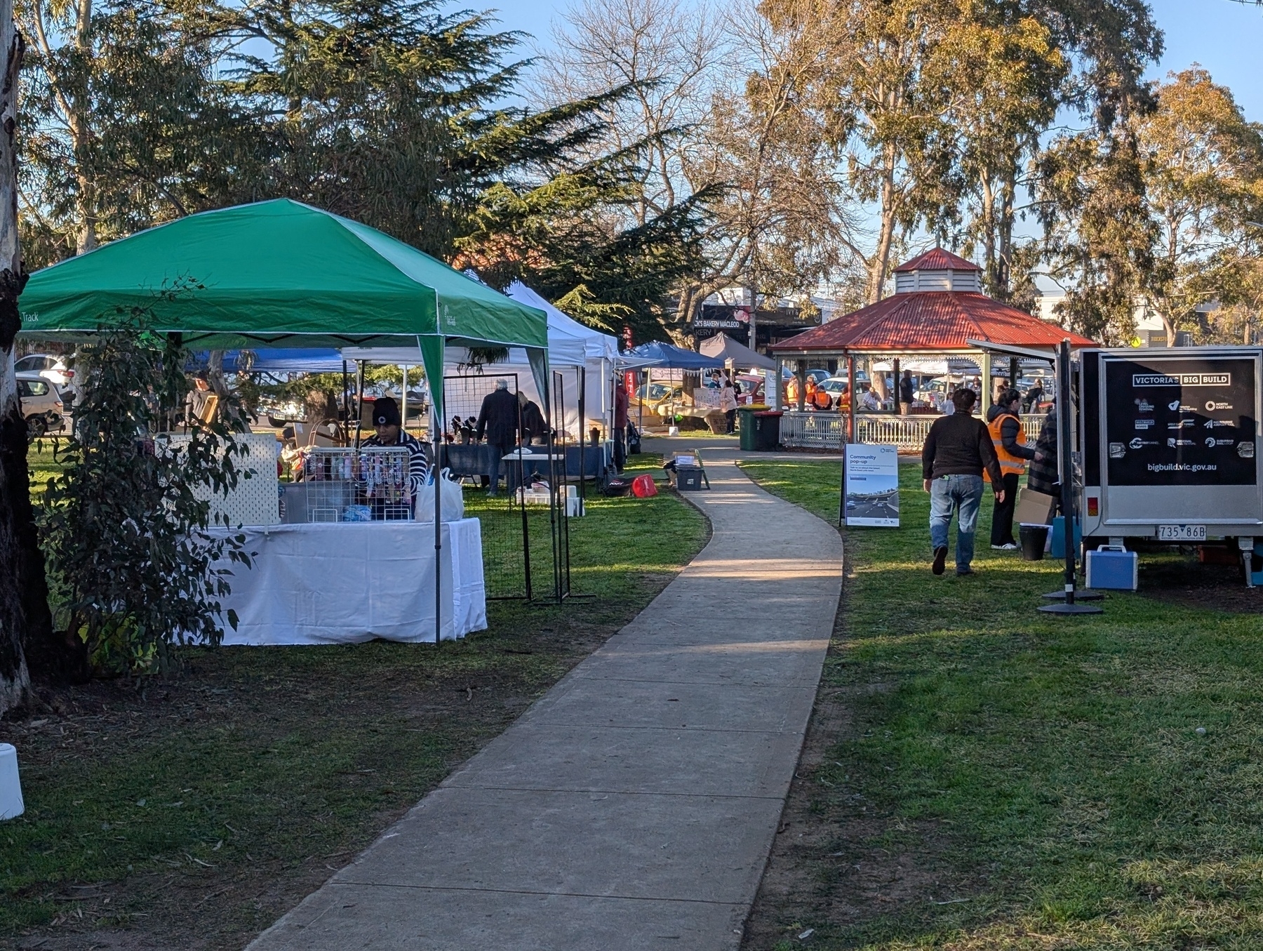Auto-generated description: A market setup in a park features various stalls with canopies along a pathway, surrounded by trees.