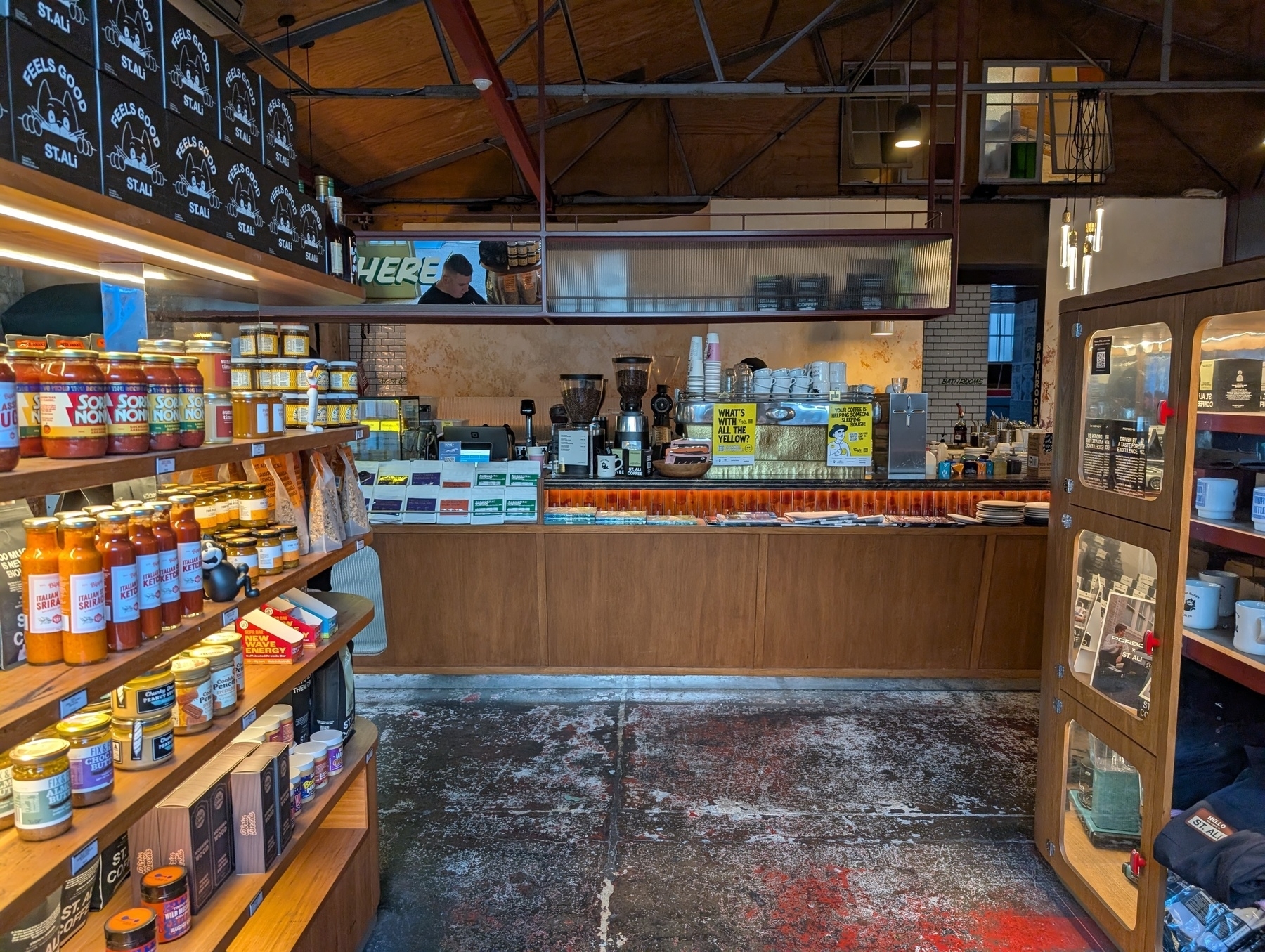 Auto-generated description: A cozy coffee shop features shelves filled with various goods, a counter with coffee-making equipment, and a warm, inviting interior.