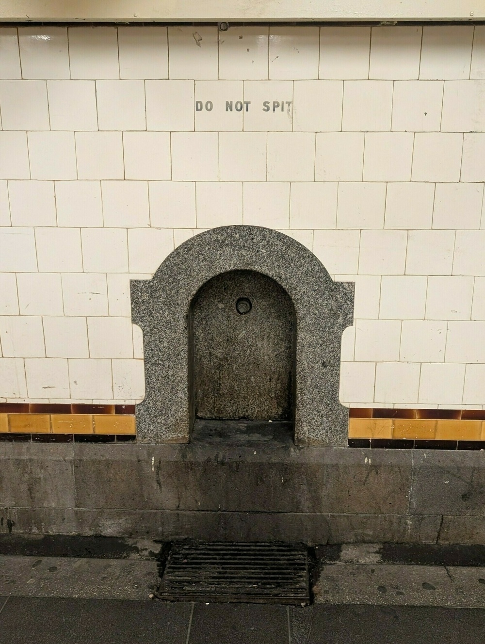 A wall-mounted drinking fountain with a sign above it that says DO NOT SPIT is set against a tiled wall.