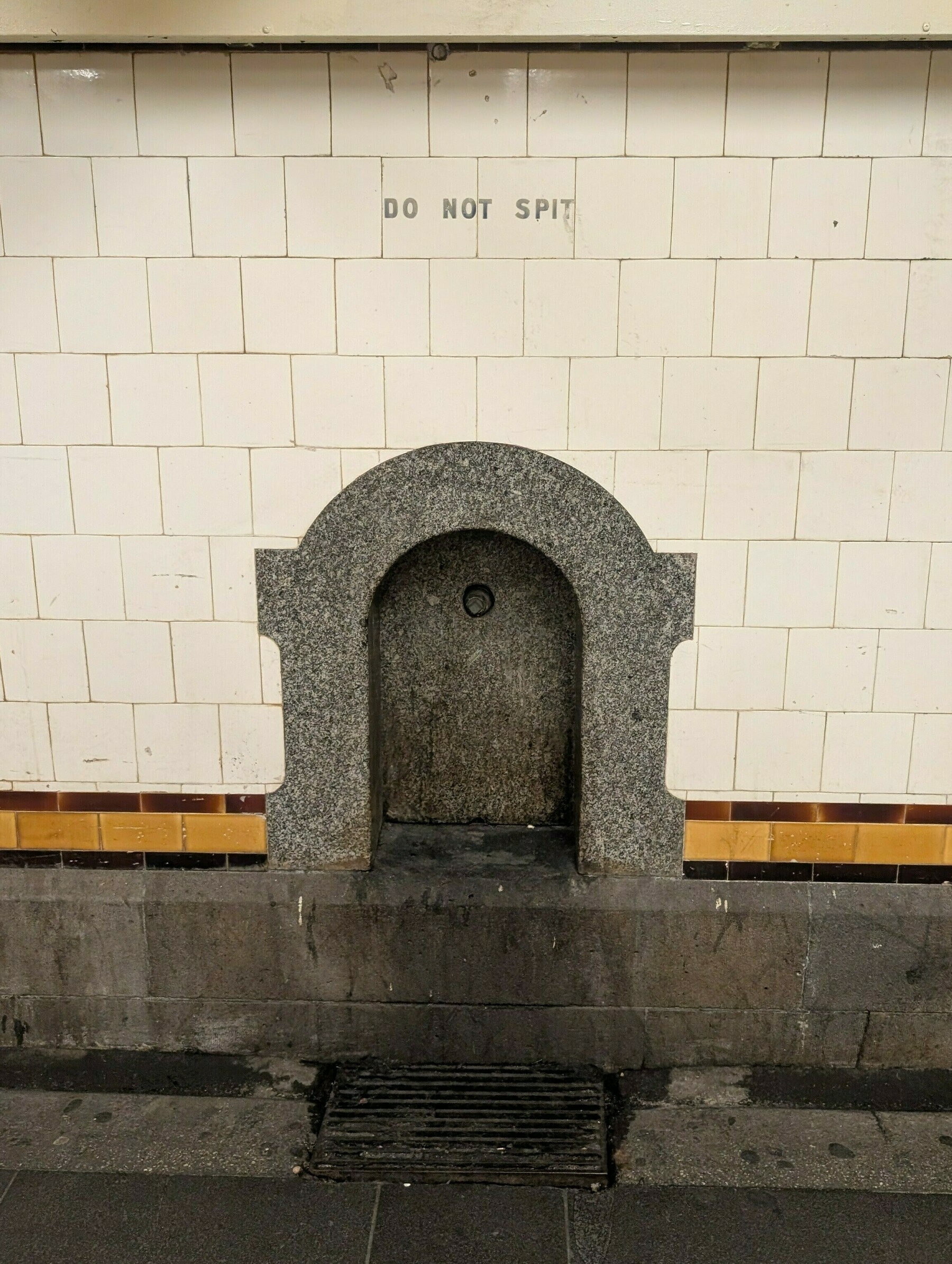 Auto-generated description: A wall-mounted drinking fountain with a sign above it that says DO NOT SPIT is set against a tiled wall.