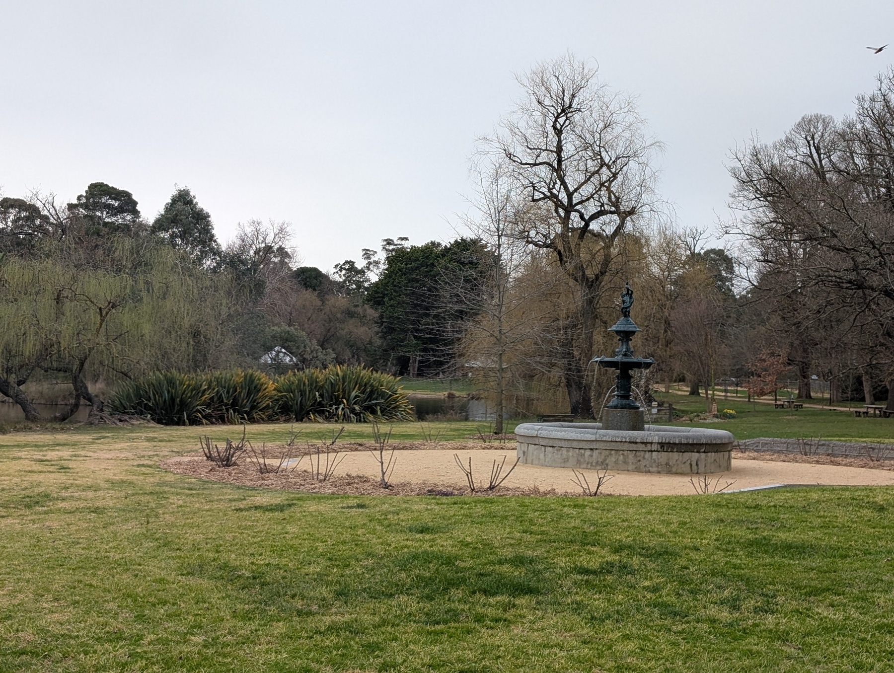 Auto-generated description: A park landscape featuring a fountain, leafless trees, and surrounding greenery under a cloudy sky.