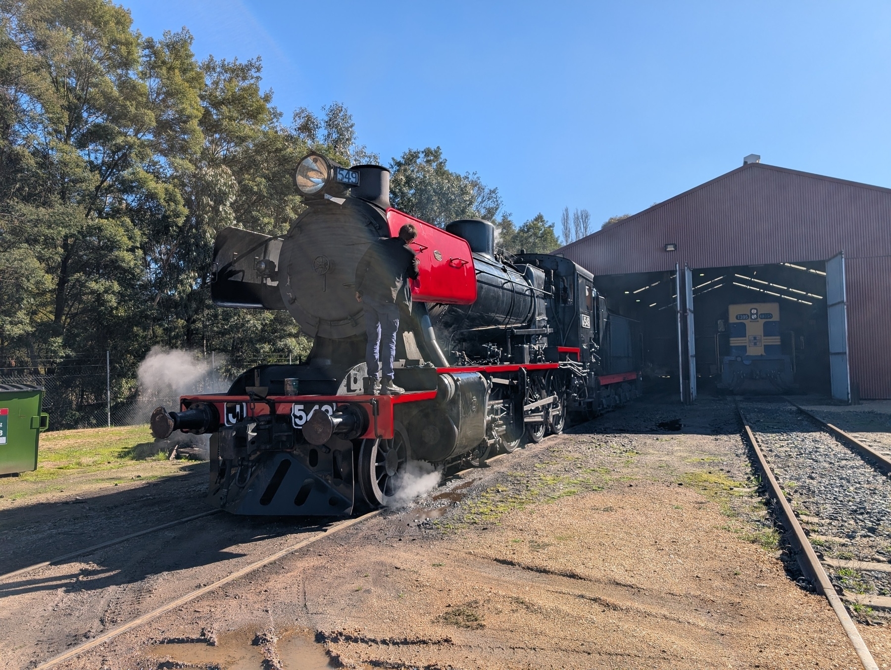 Auto-generated description: A steam locomotive with a red front is exiting a shed on a sunny day.