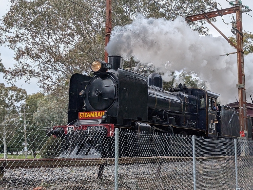 A vintage steam locomotive is producing white smoke while traveling on train tracks behind a chain-link fence.