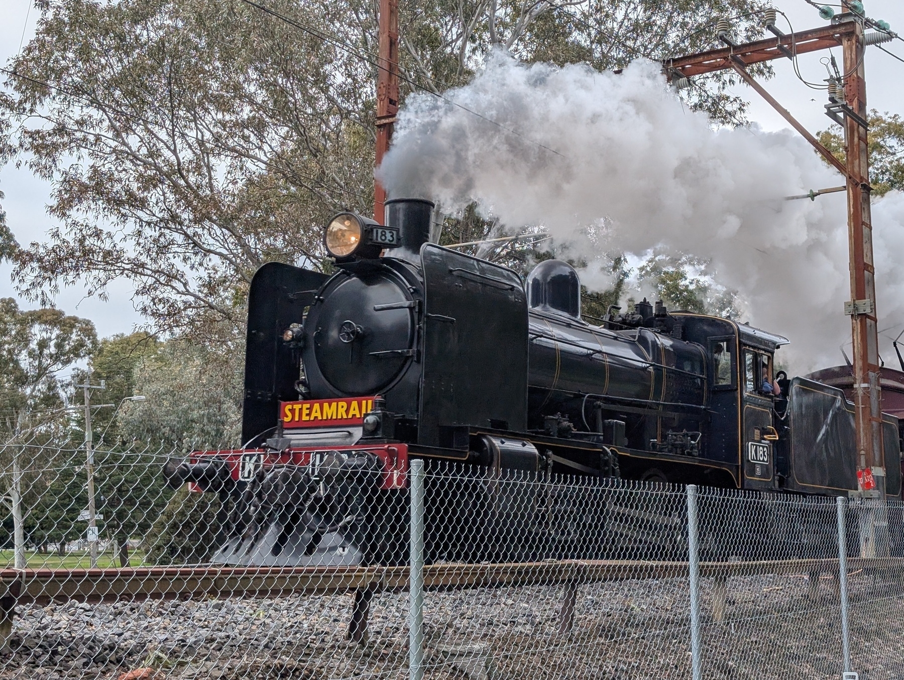 Auto-generated description: A vintage steam locomotive is producing white smoke while traveling on train tracks behind a chain-link fence.