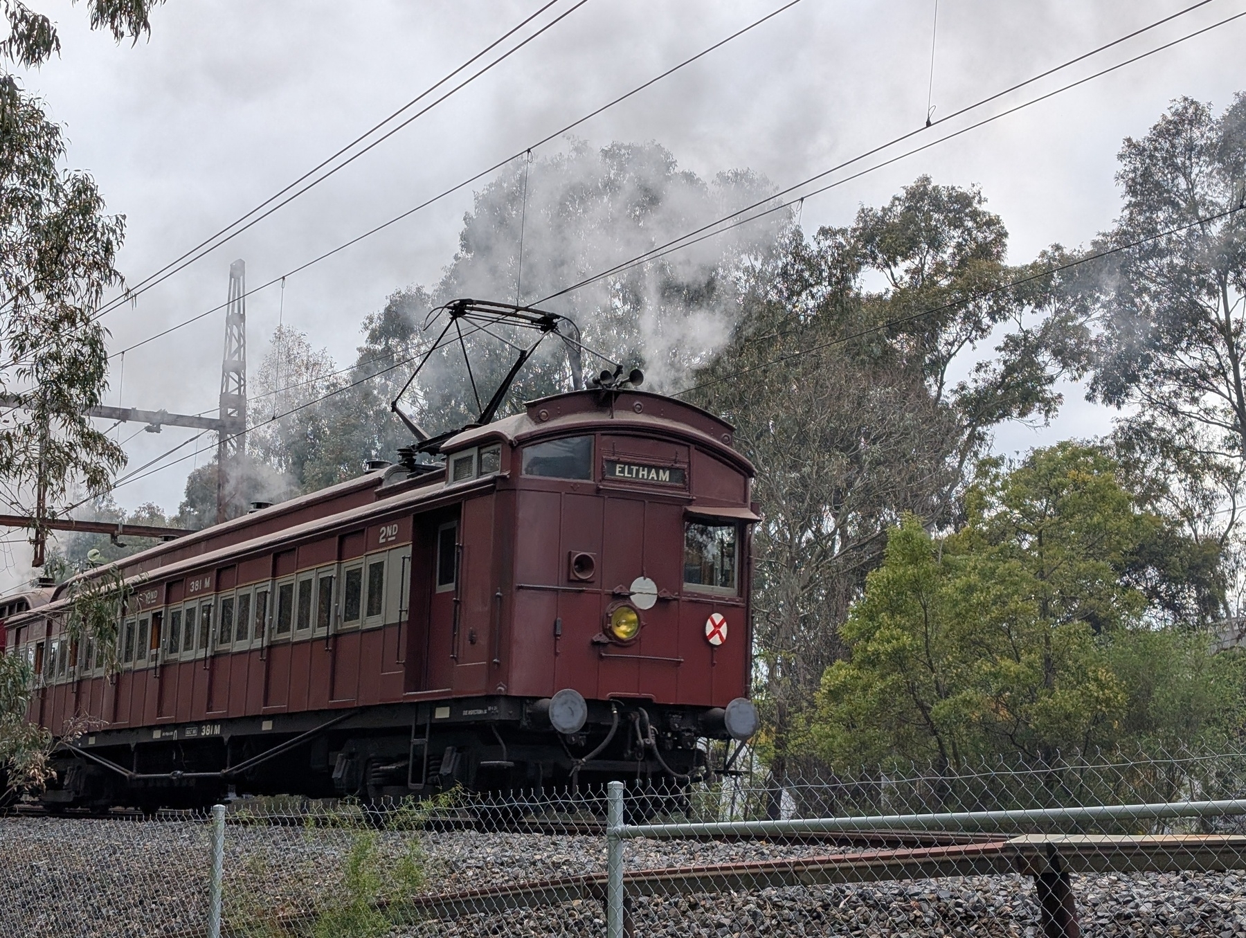 Auto-generated description: A vintage red Tait train labeled Eltham is moving along railway tracks surrounded by trees and emitting white smoke.