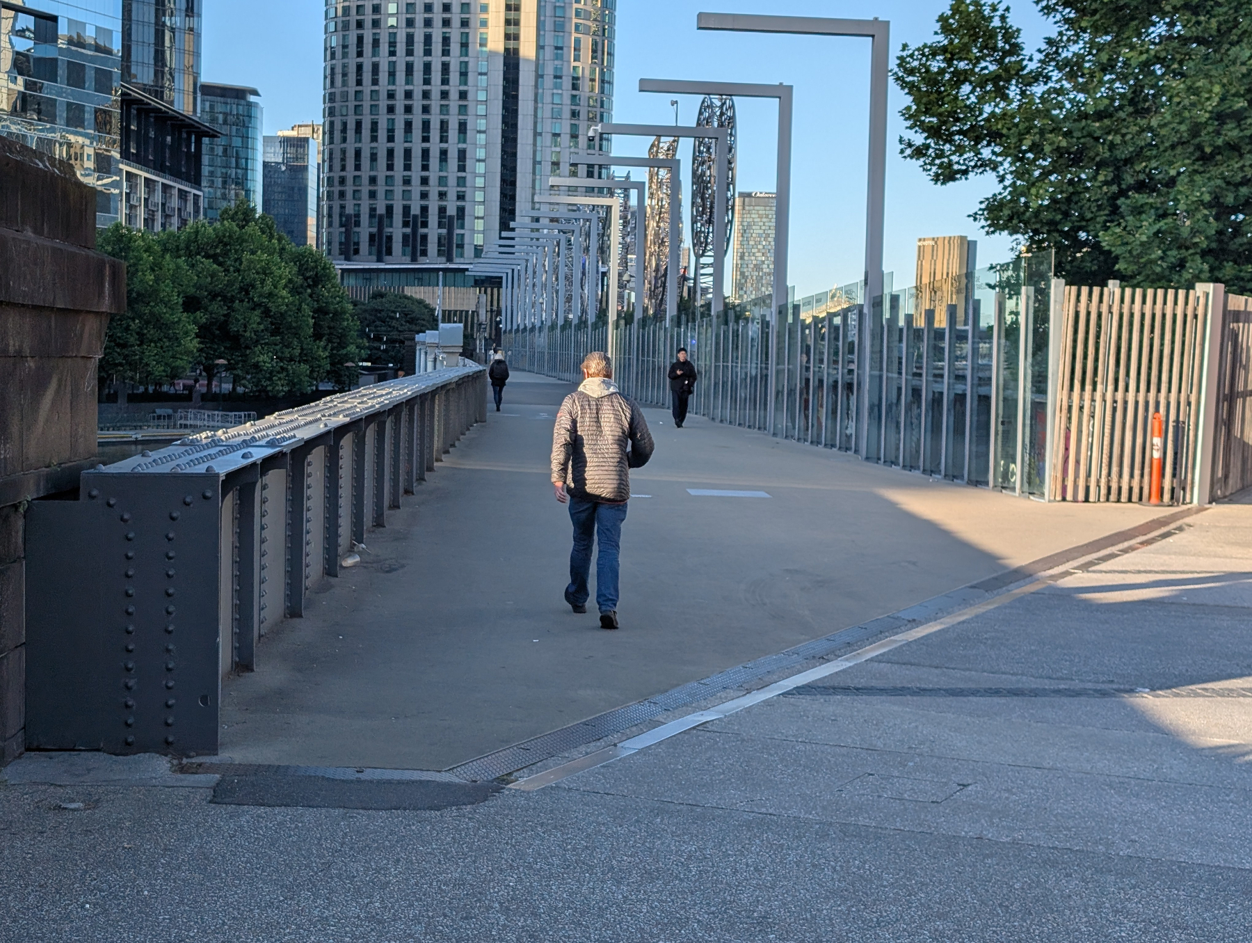 A person is walking across a bridge that branches from a path lined with trees and modern buildings in the background.