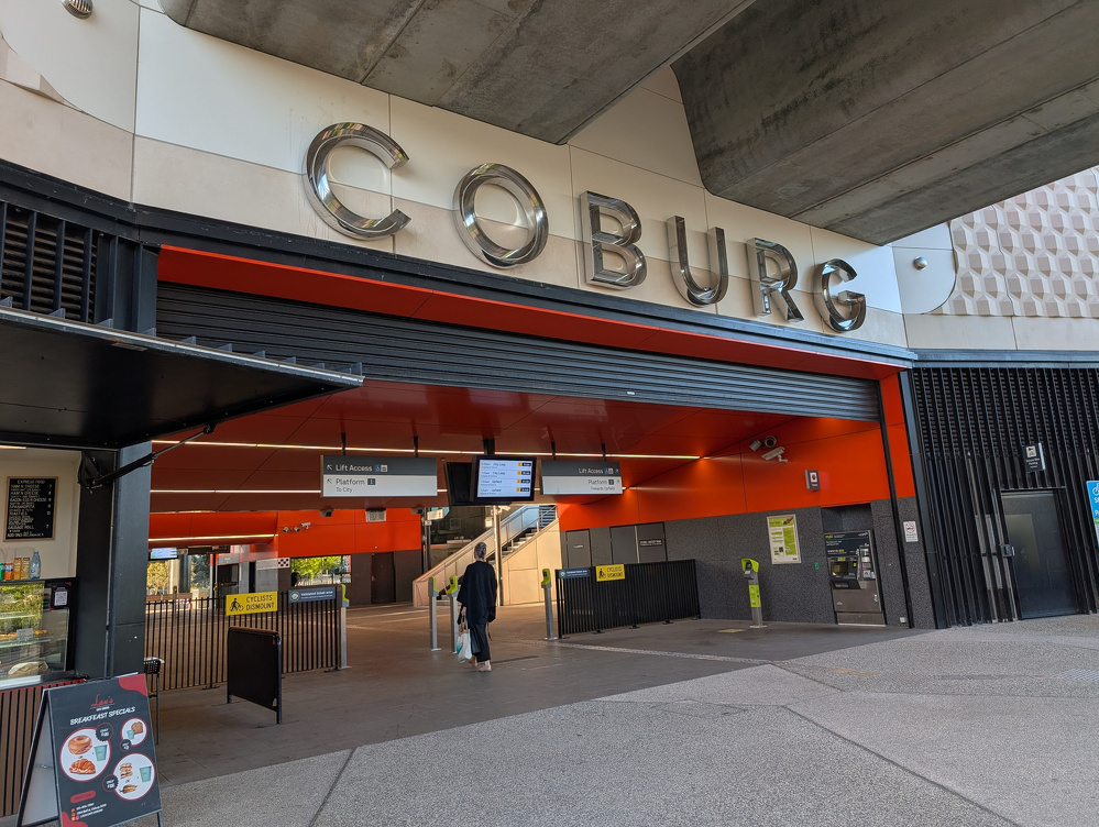 A person walks towards the entrance of a modern train station labeled Coburg with various signs and a small food stand nearby.