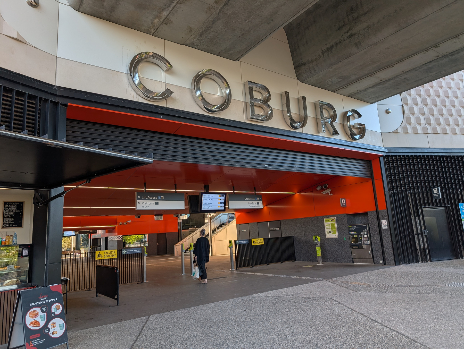 Auto-generated description: A person walks towards the entrance of a modern train station labeled Coburg with various signs and a small food stand nearby.