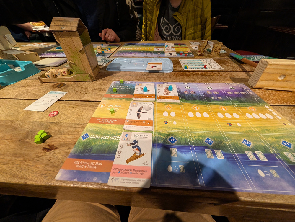 A group of people are gathered around a wooden table playing a board game with various colorful cards and pieces.