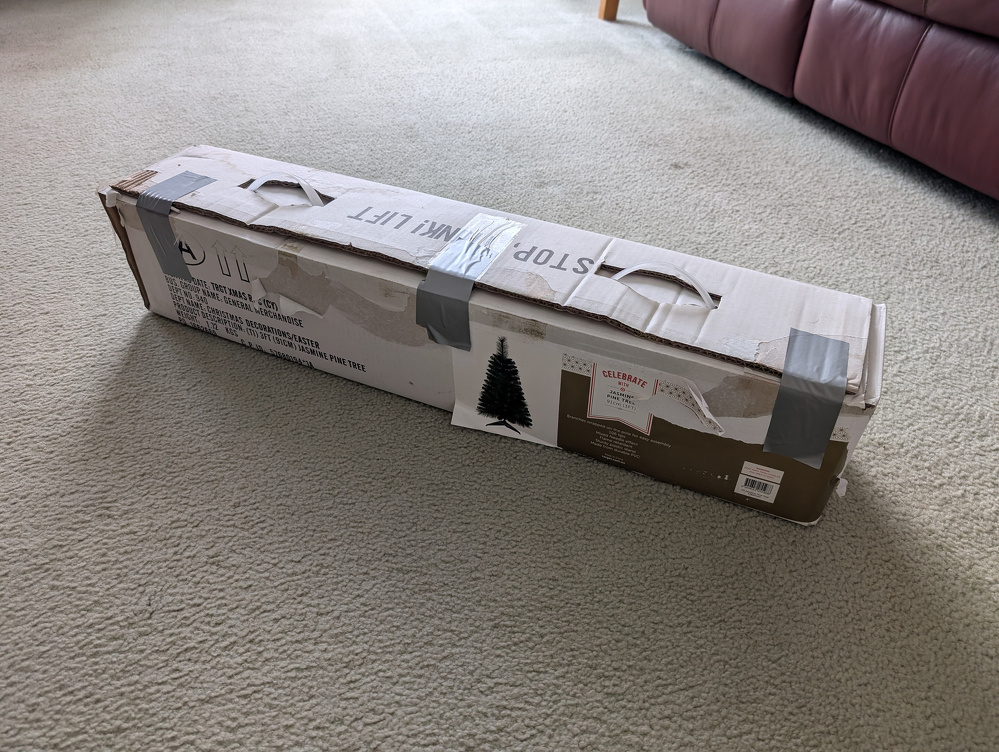 A closed cardboard box containing an artificial Christmas tree is sealed with tape and placed on a carpeted floor beside a leather sofa.
