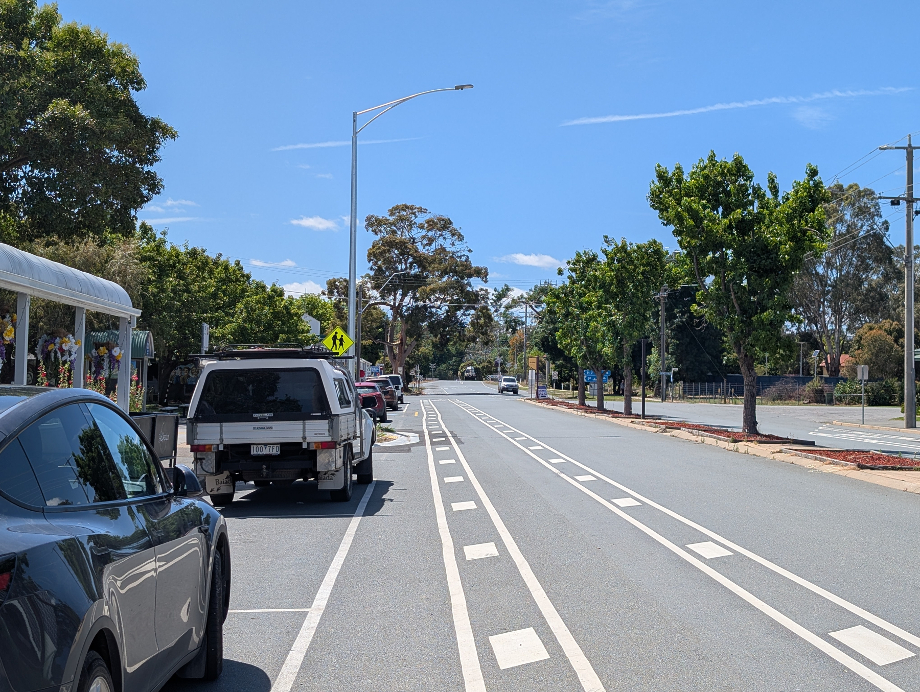 Auto-generated description: A quiet street features parked cars, bicycle lanes, clear skies, and leafy trees lining the road.