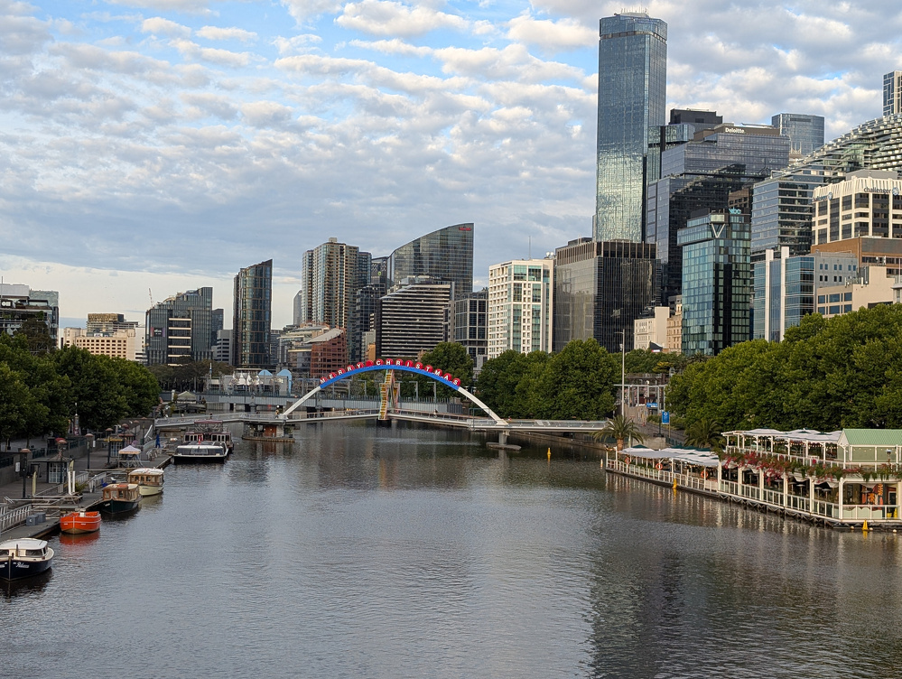 A modern city skyline with a river running through it features a pedestrian bridge and numerous high-rise buildings.