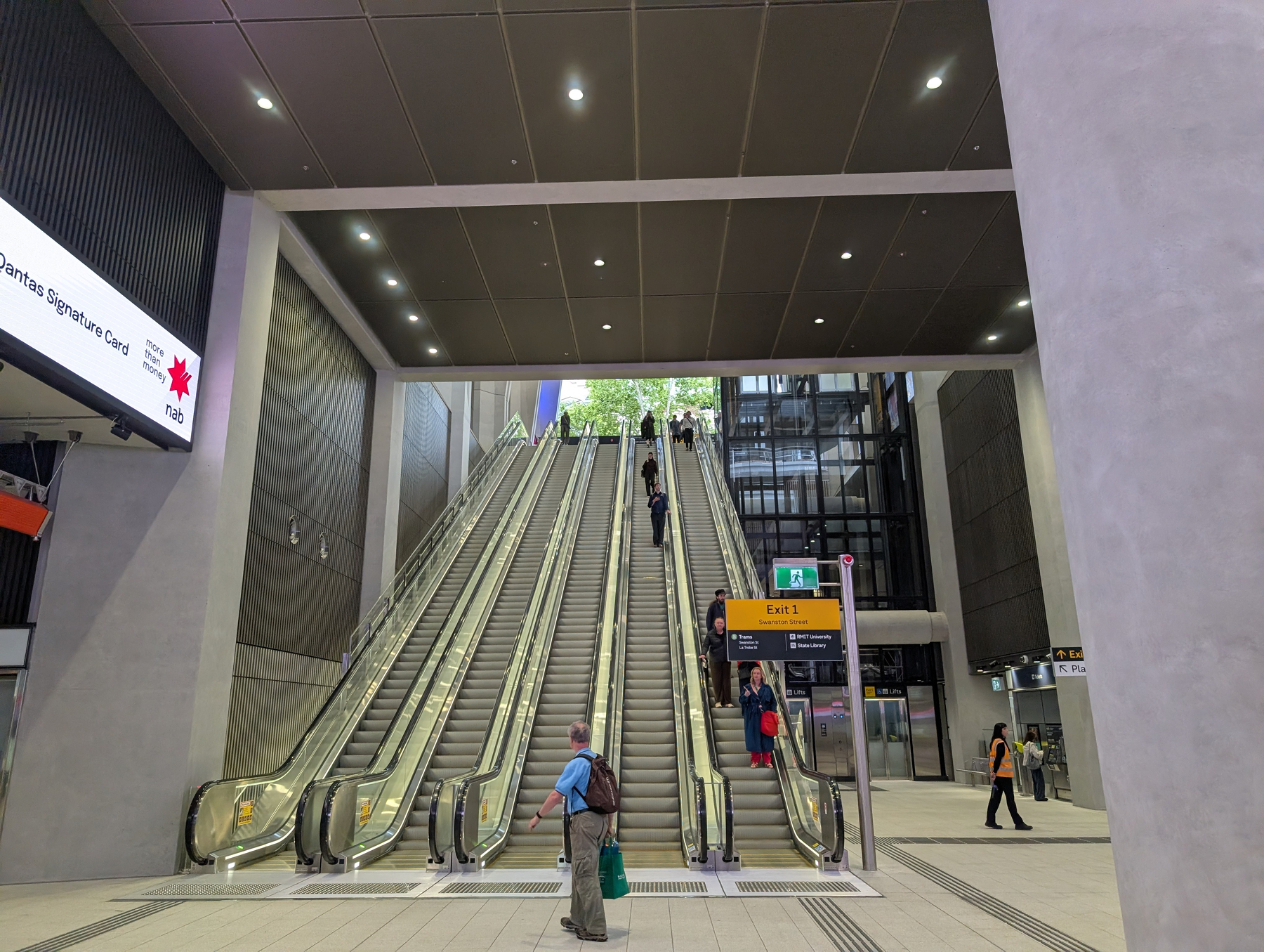 Auto-generated description: An indoor view of a modern transit station features a long escalator with people ascending and overhead signs guiding commuters.