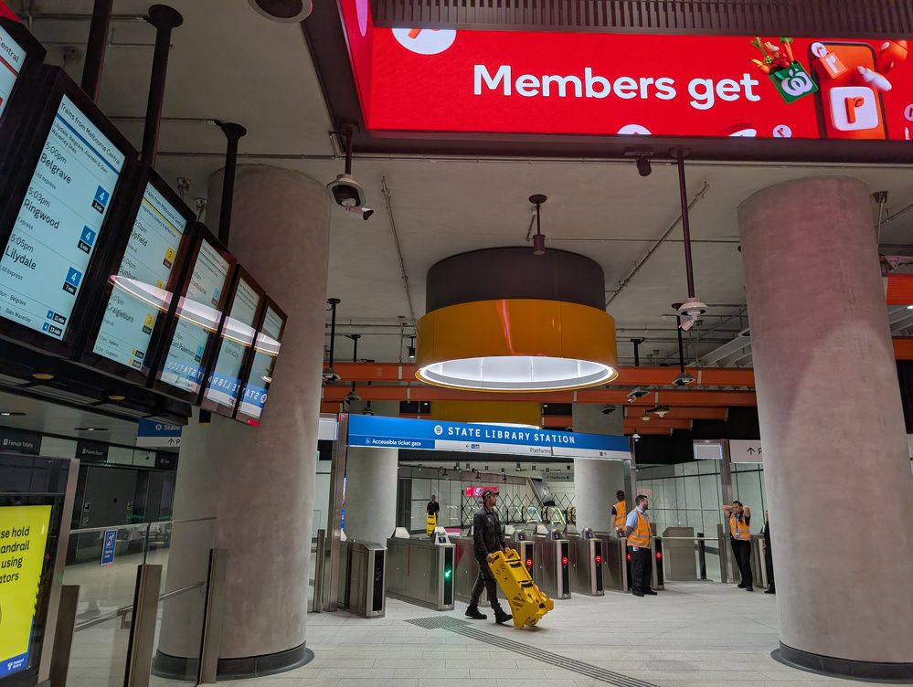 A modern train station features digital screens, advertisements, and passengers near ticket barriers.