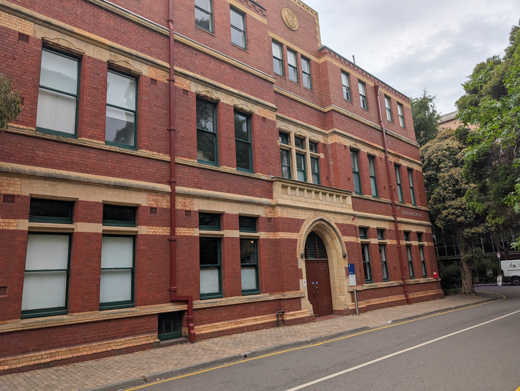Auto-generated description: A red brick building with multiple windows and an arched entrance stands along a quiet, tree-lined street.