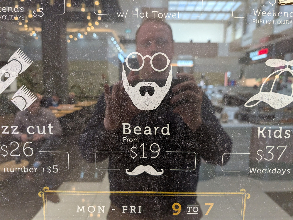 A window advertisement for barber services, featuring prices for buzz cuts and beard grooming, along with reflection of a person taking a photo.