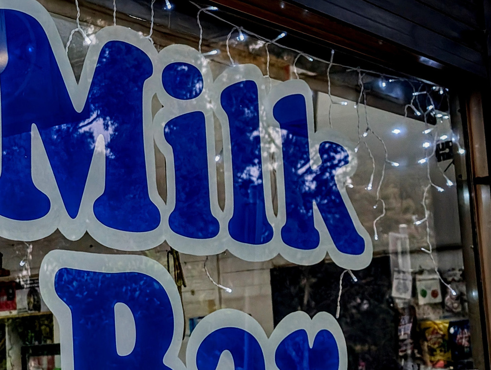 Large blue lettering spelling Milk Bar is displayed on a window adorned with string lights.